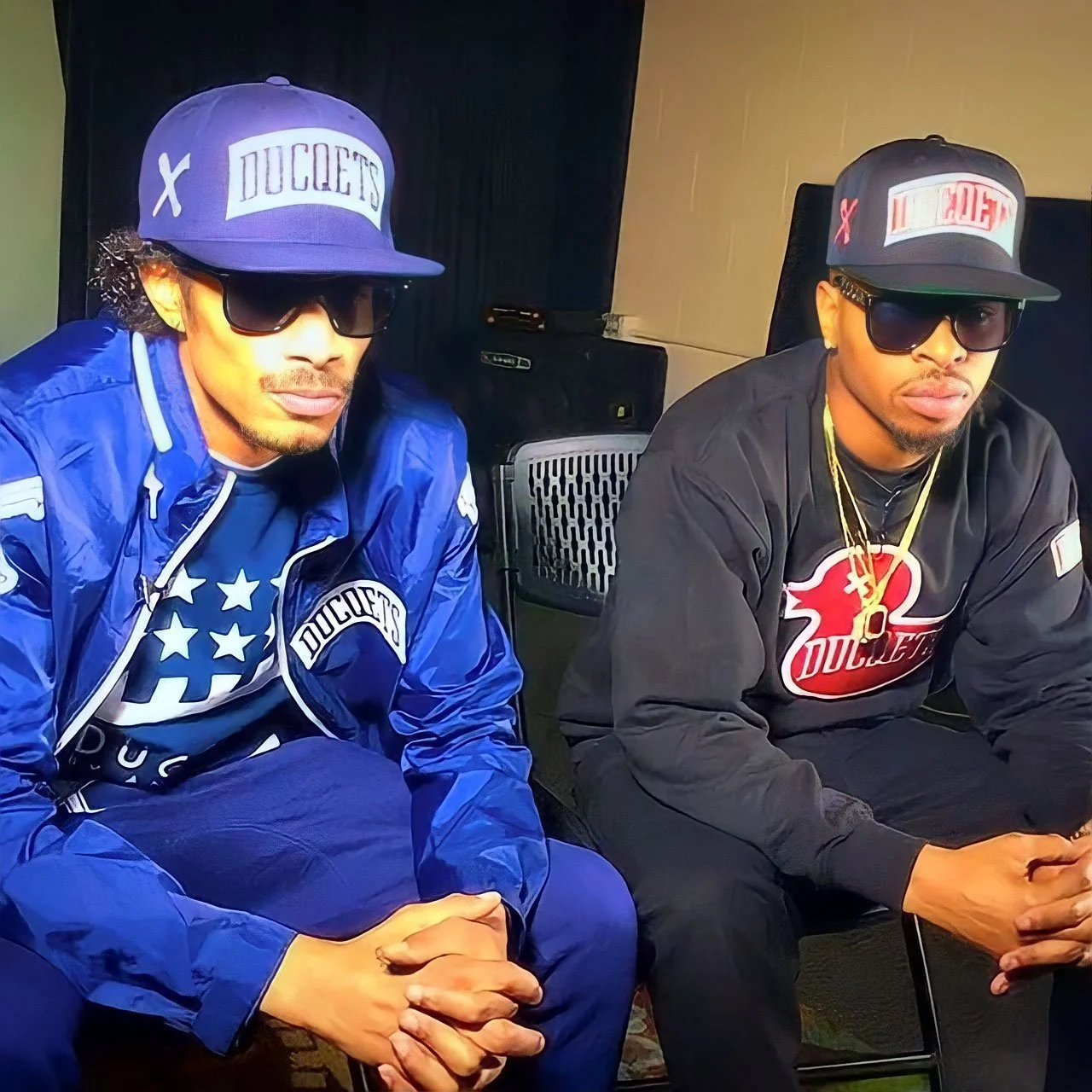 Two men wearing baseball caps, sunglasses, and black clothing, seated indoors with sound equipment in the background.
