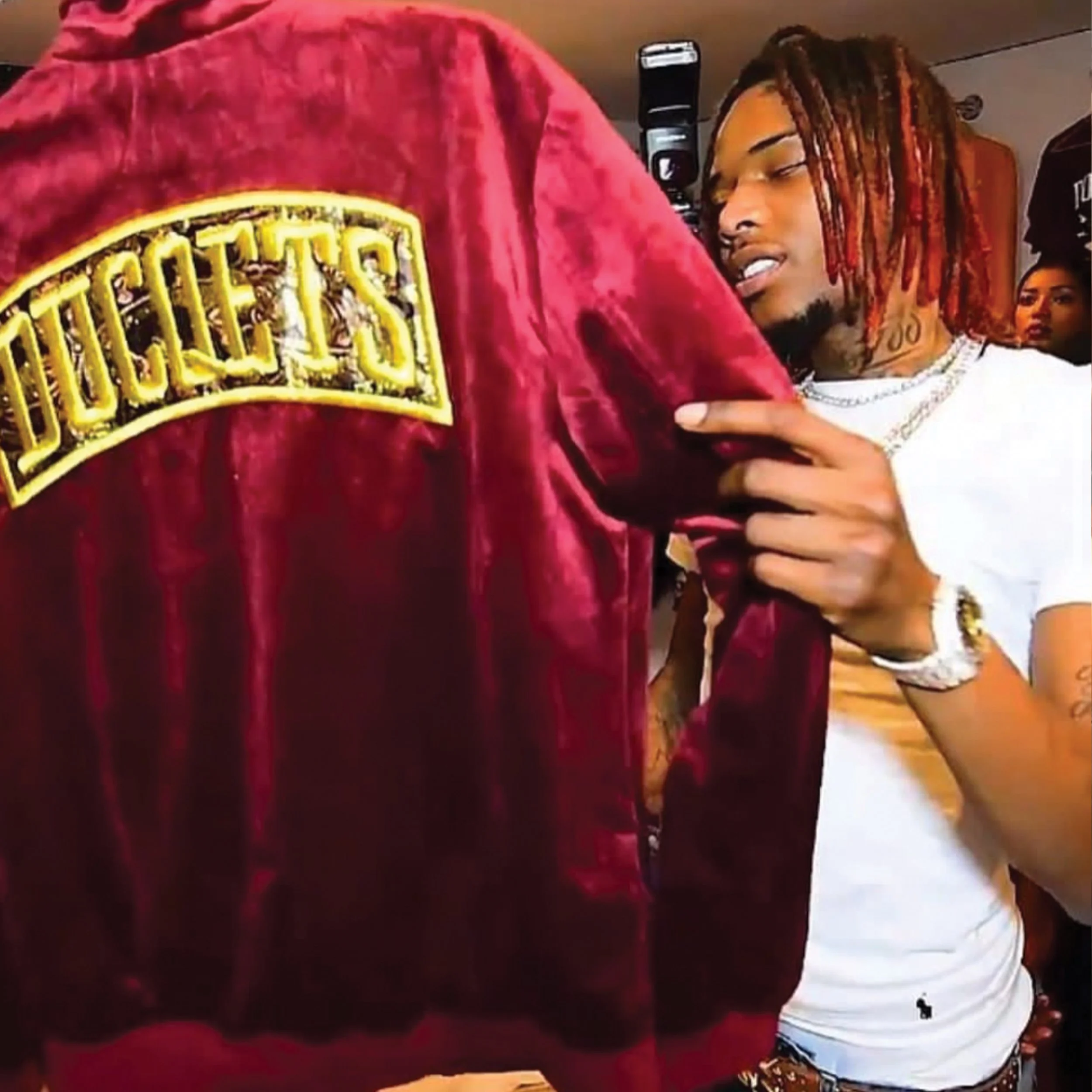 A young man with orange dreadlocks, a tattoo on his neck, wearing a white T-shirt, pointing to a person in a red jacket with a large yellow embroidered patch on the back that says "TOUCHE". The young man appears to be engaged in conversation or inter