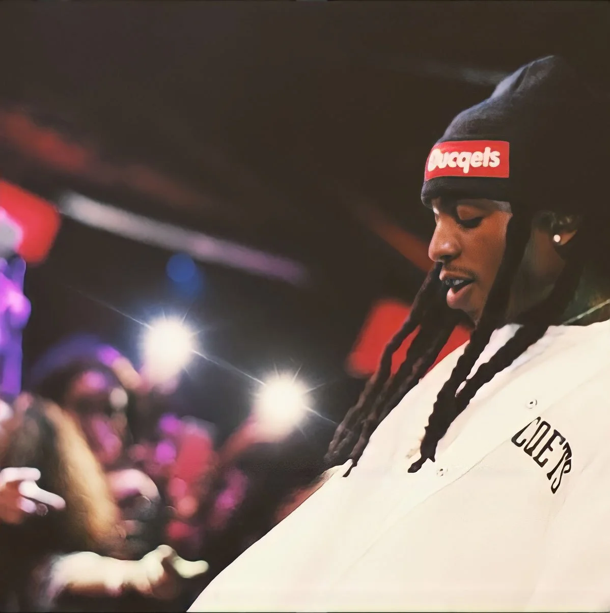 A young man with long braids wearing a black beanie with a red "Nuggets" logo, and a white coat with "COLE