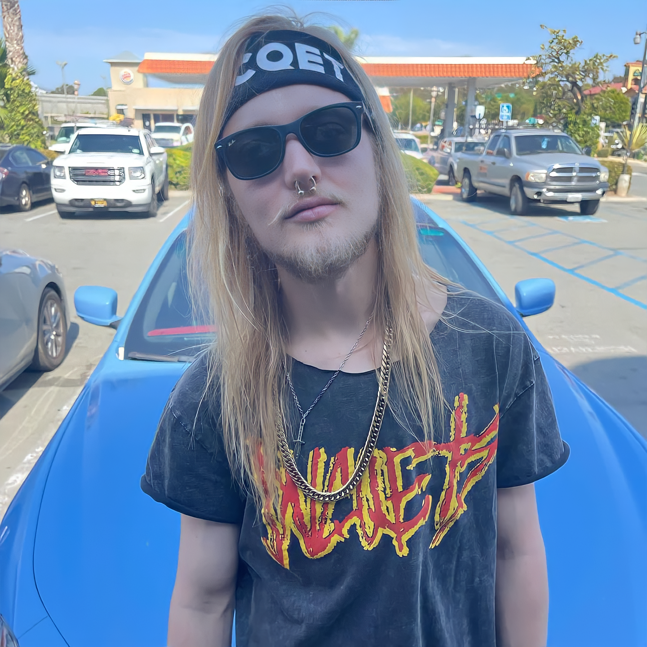 A young man with long blond hair, wearing black sunglasses, a headband, and a septum piercing, stands in front of a blue car in a parking lot, wearing a black graphic T-shirt with yellow and red lettering.