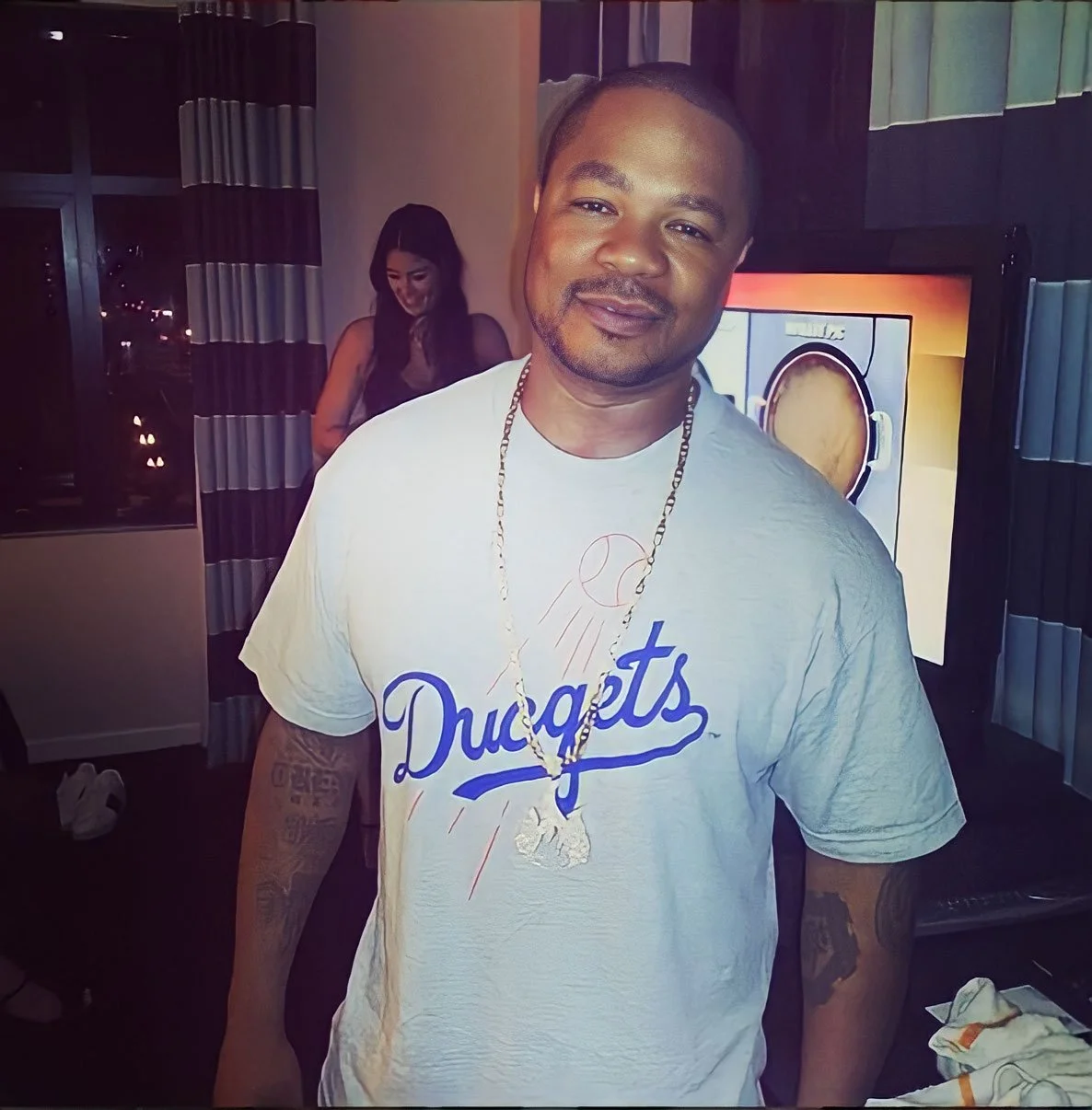 A man with short hair and a goatee, wearing a white T-shirt with 'Dugat' written on it and a gold chain, is smiling and standing indoors. A woman in a black top is in the background, looking down. The room has black and white curtains, a television d