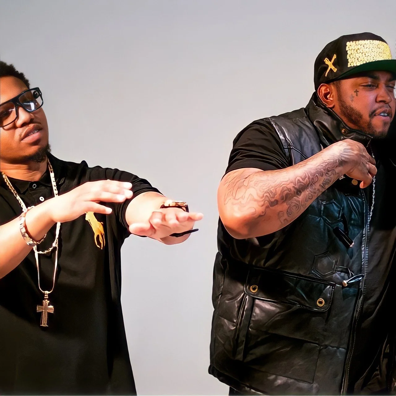 Two men with tattoos and jewelry, one with glasses, gesturing with their hands against a plain light background.