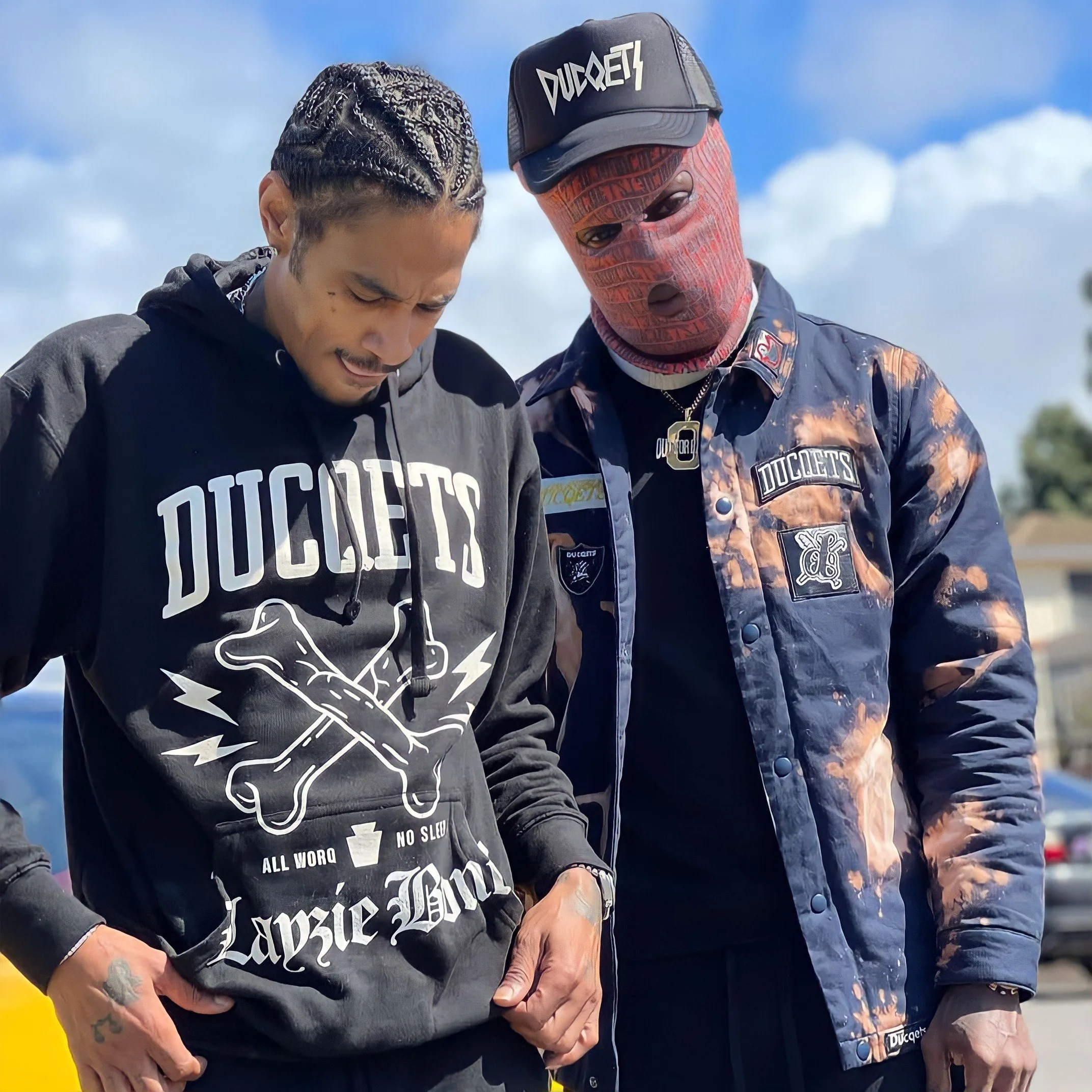 Two young men standing outdoors, looking down, wearing dark streetwear with logos and patches, against a blue sky with some clouds.