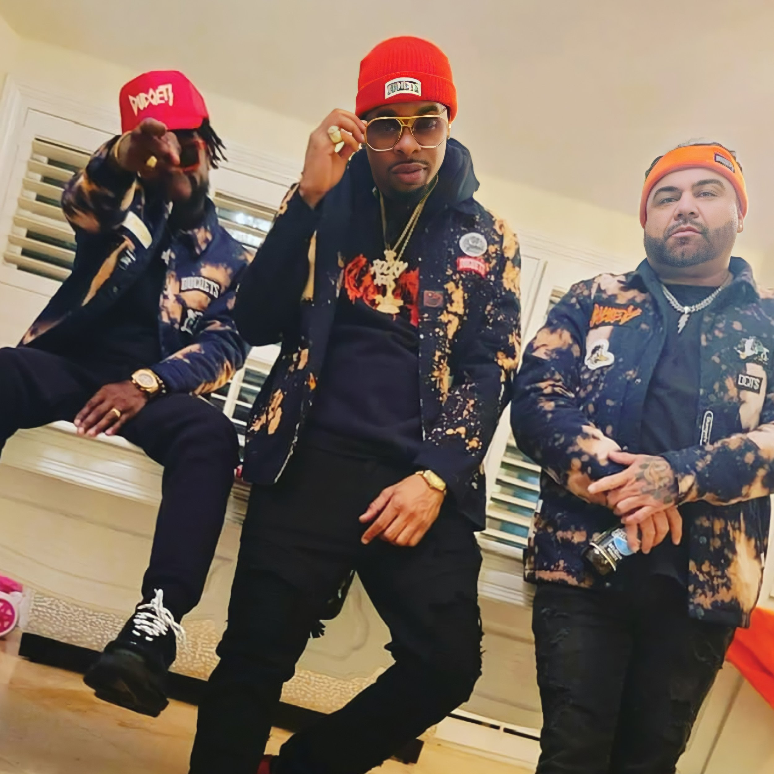 Three men dressed in black and gold patterned jackets, some wearing orange or red beanies, and accessories like sunglasses, necklaces, and watches, posing indoors.