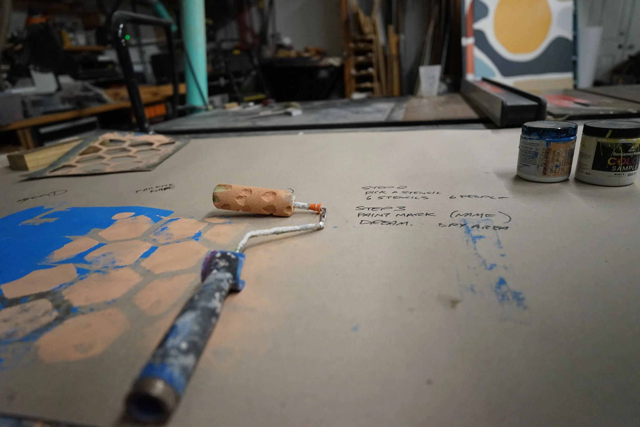 Work table with a paint roller, painted sheet with blue and beige patterns, and two containers of paint. Handwritten notes on the table indicate steps for creating art.