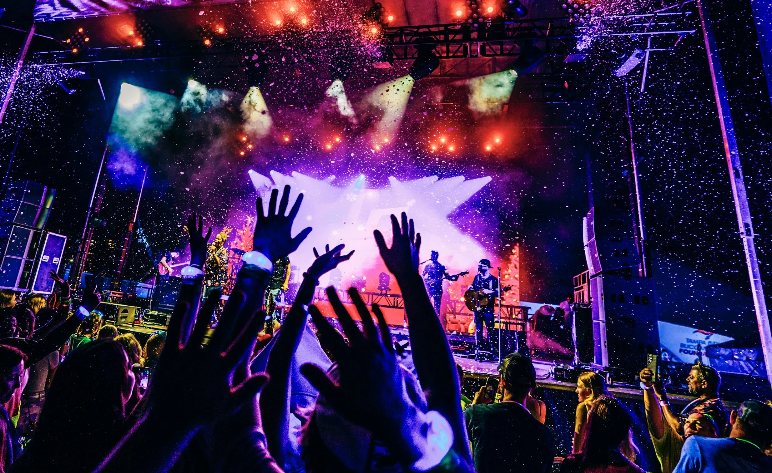 A concert stage with colorful lights, smoke, and confetti. Hands of the audience raised, some with glow bracelets, as musicians perform.