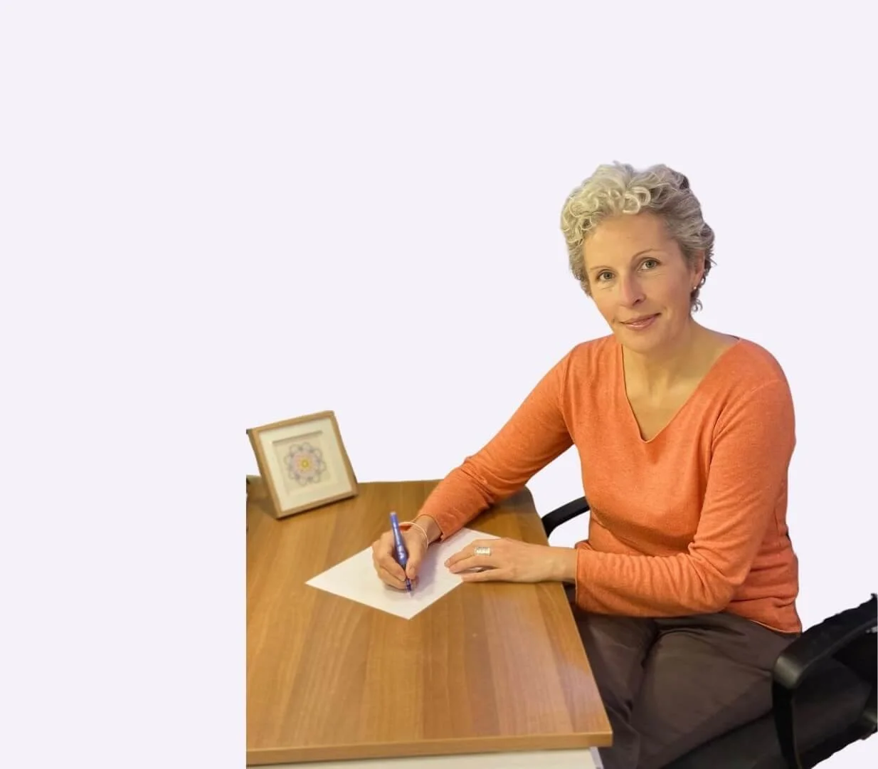 A woman with short curly gray hair, wearing an orange long-sleeve shirt, sits at a wooden desk writing on a sheet of paper with a blue pen. There is a small framed piece of artwork on the desk and a black chair.
