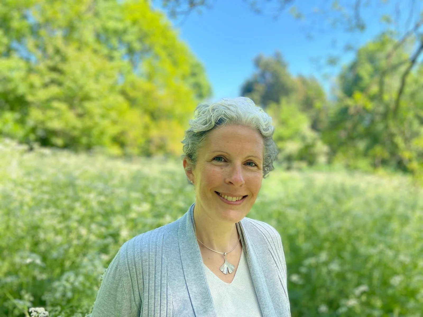 A woman with short, curly gray hair smiling outdoors in a green park with trees and blue sky in the background.