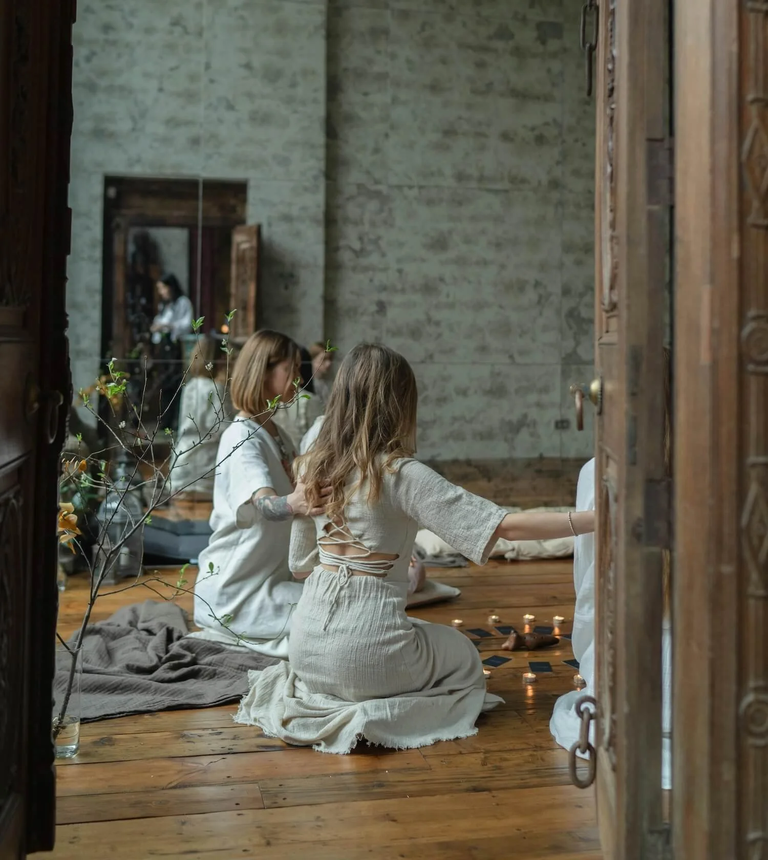 Three women with light skin and brown hair sitting on a wooden floor, engaged in a spiritual or meditative ritual, surrounded by candles and plant branches in a rustic room with textured gray walls and large mirror.