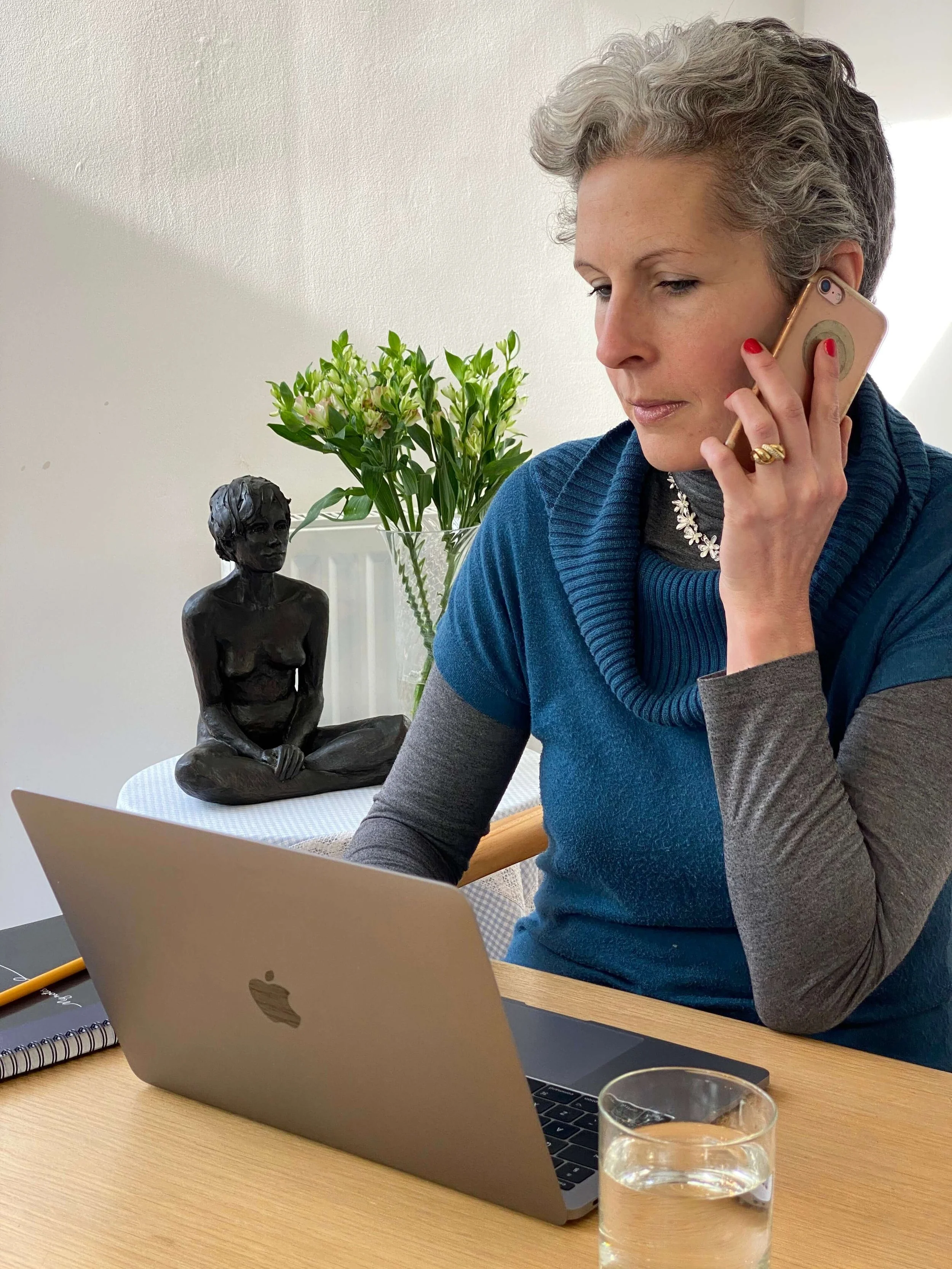 A woman with short gray hair wearing a blue and gray long-sleeve shirt, talking on a cell phone, seated at a wooden table with a MacBook laptop, a glass of water, a notebook, and a pen. Behind her, a black statue of a seated woman and a vase with green flowers are visible.