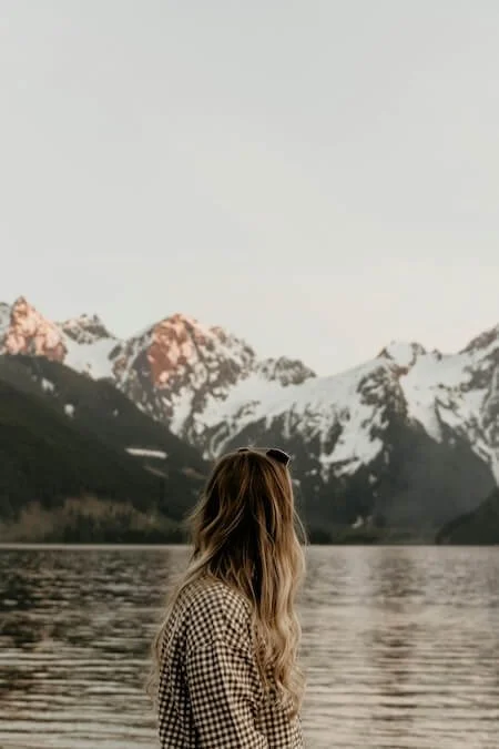 A woman with long, wavy hair standing by a lake, looking at snow-capped mountains during sunset.
