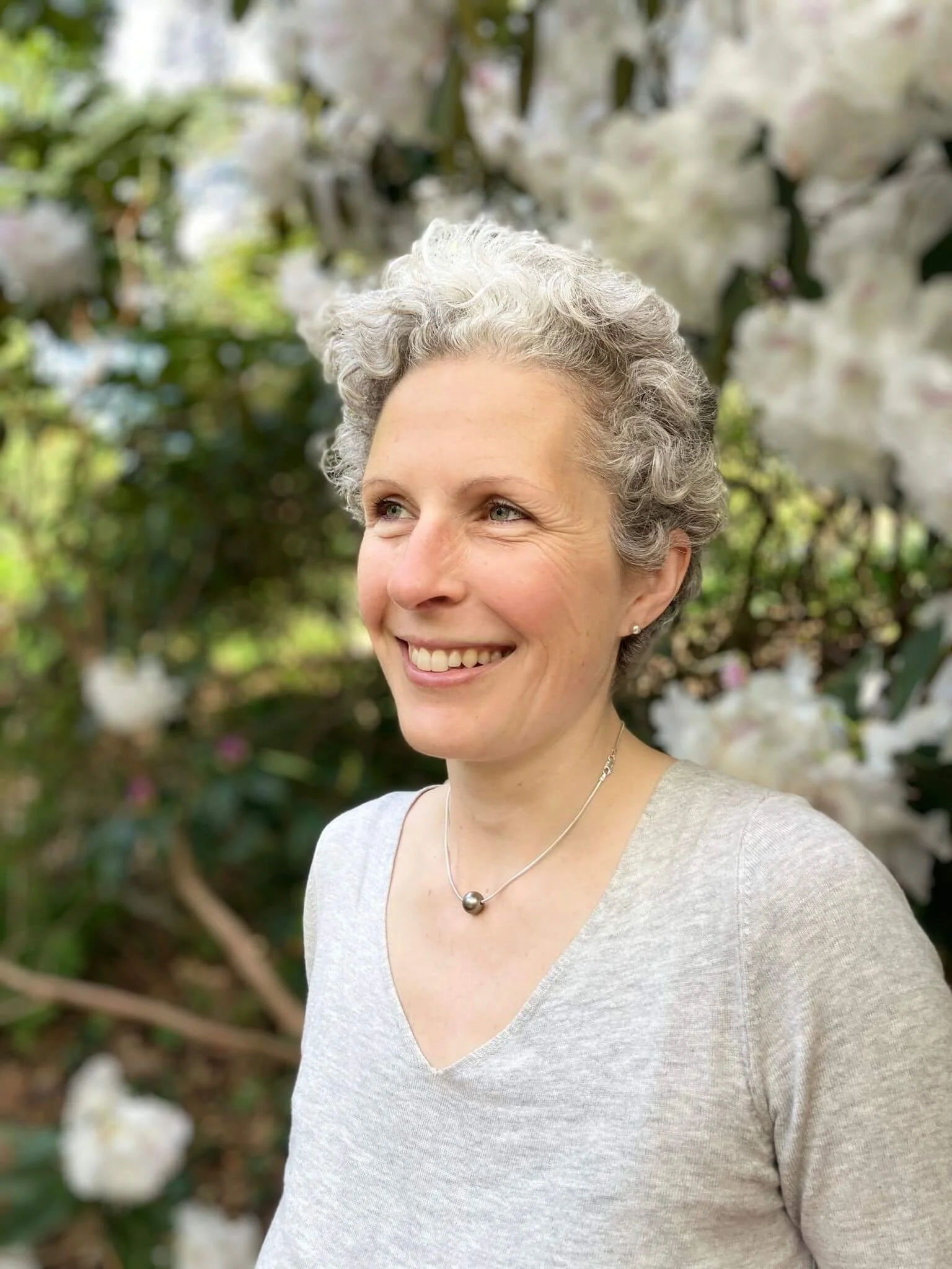 A woman with short, curly gray hair smiling outdoors in front of blooming white flowers and green foliage.