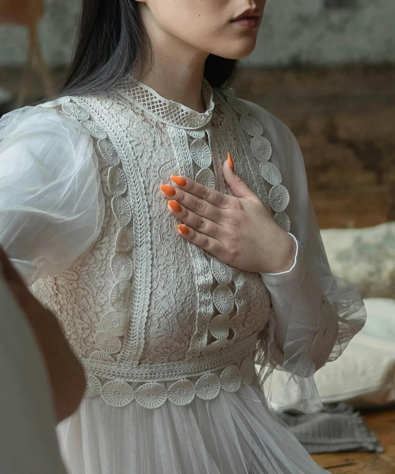 A woman wearing a vintage white lace dress with intricate embroidery, holding her hand over her chest, showing orange-painted nails.
