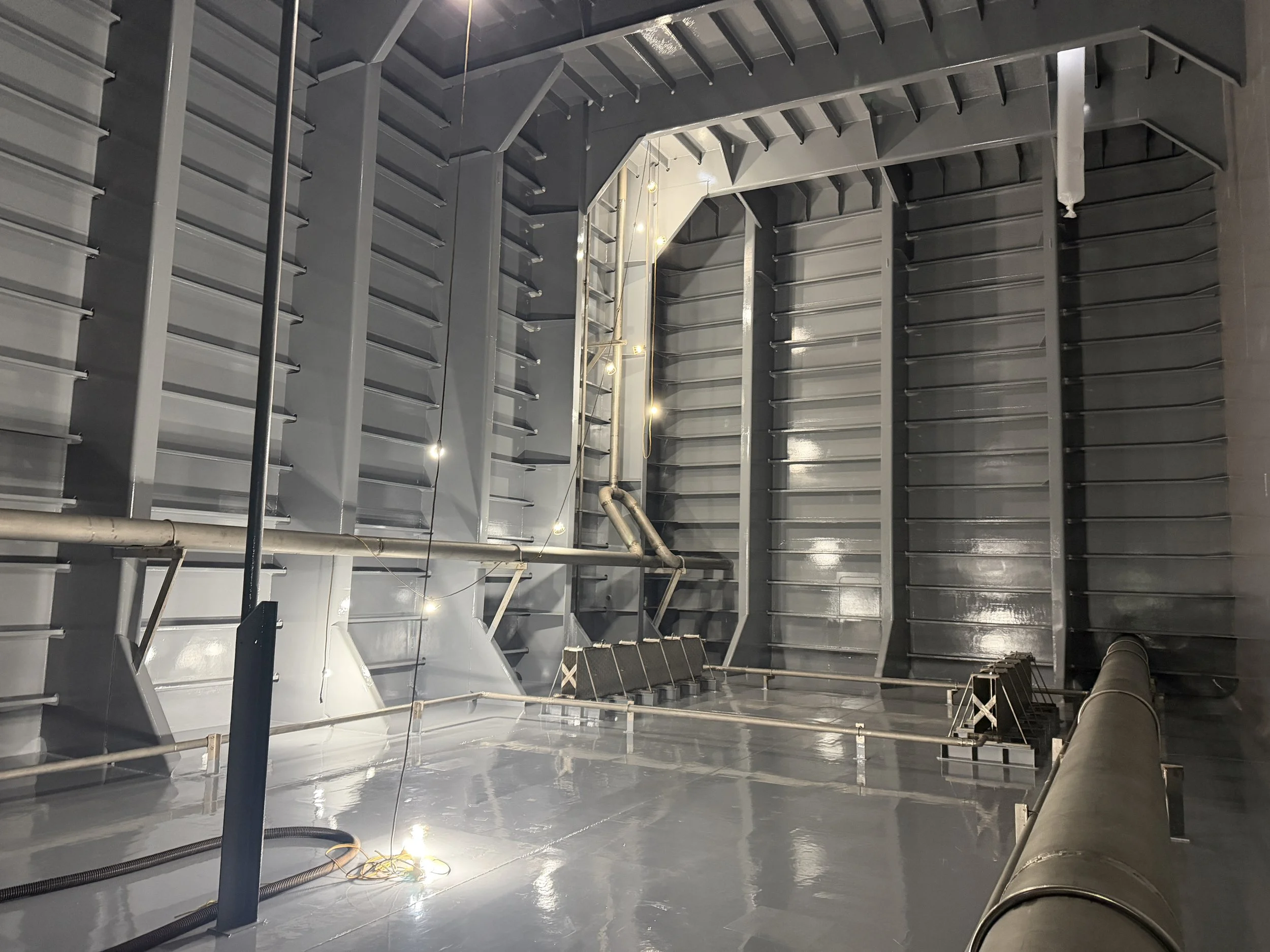 Interior of an industrial metal tank with ribbed walls, pipes, structural supports, and temporary work lights