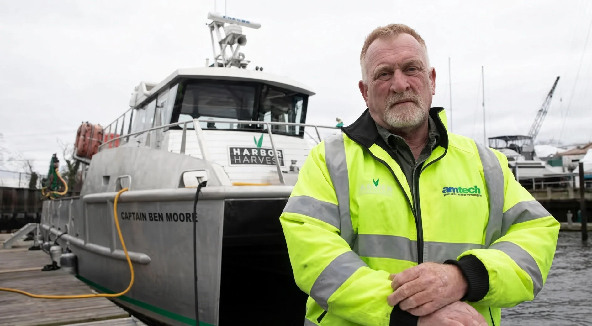 Man in a high-visibility safety jacket standing beside the Harbor Harvest boat Captain Ben Moore at a marina