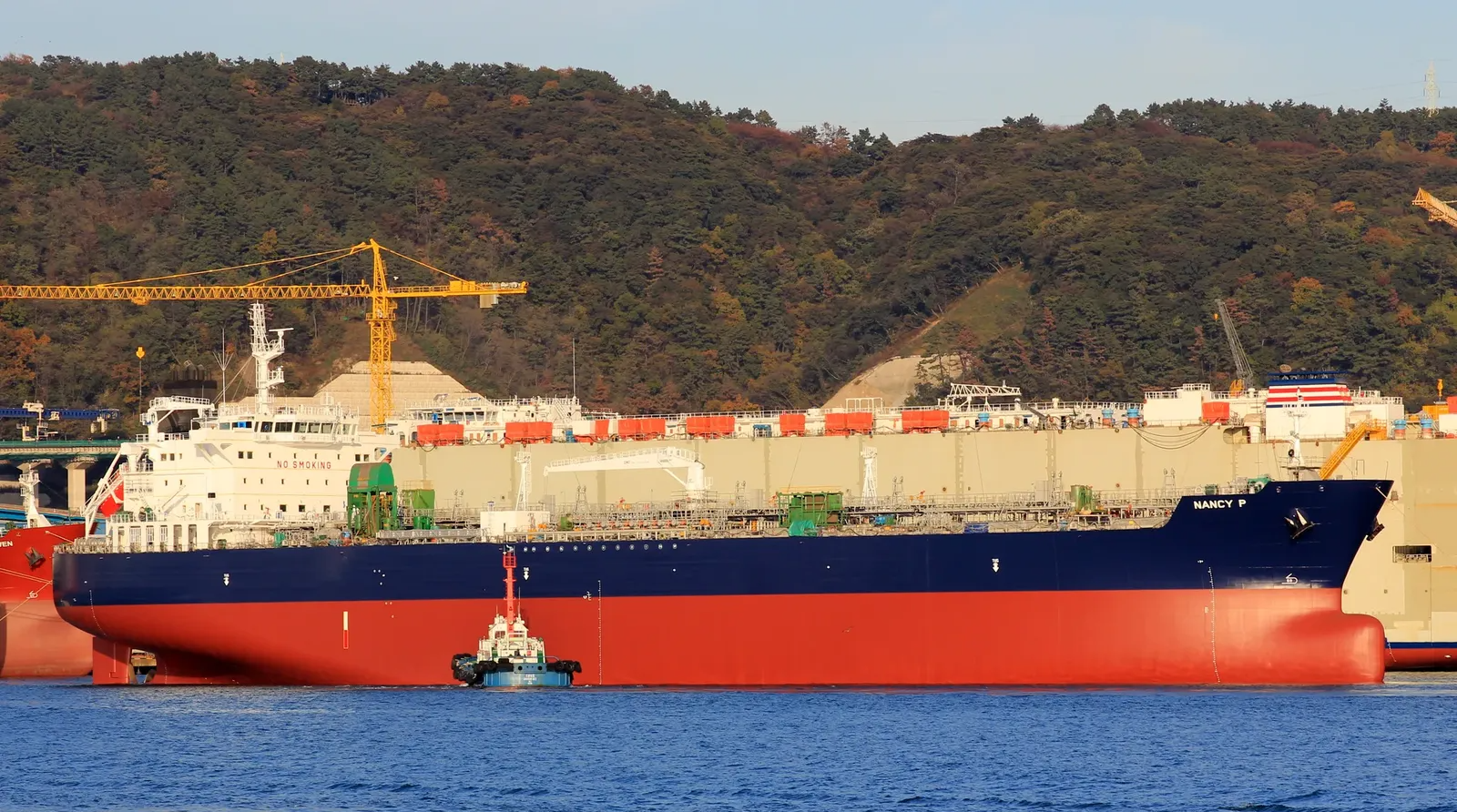 Large red-and-navy cargo ship named Nancy P docked beside a tugboat, with yellow construction cranes and an autumn-covered hillside in the background