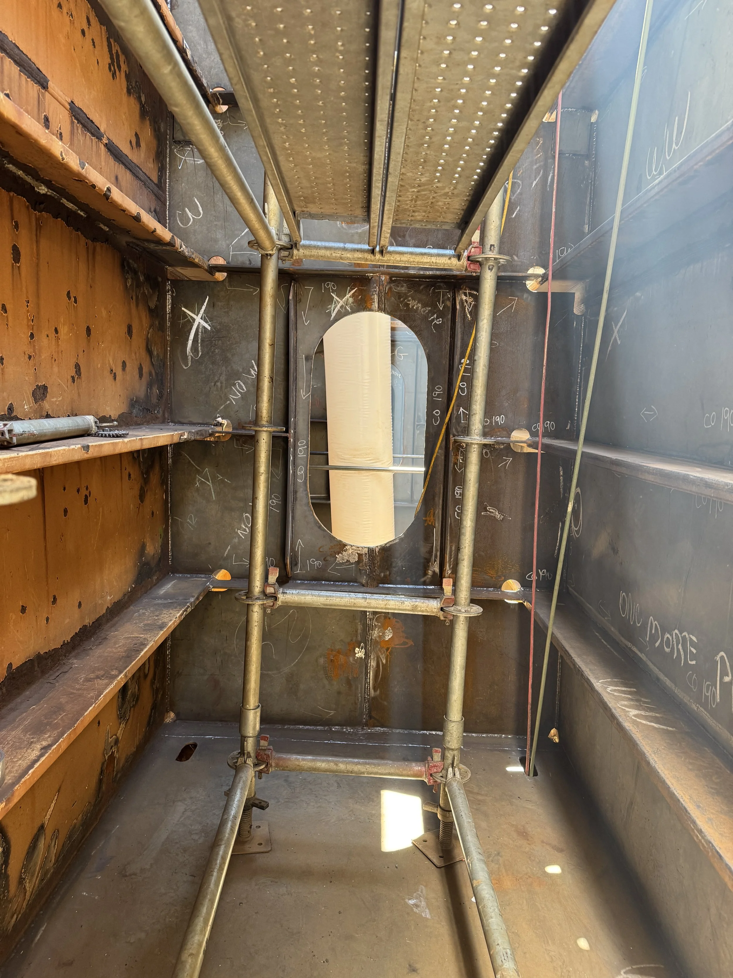 Interior of a steel industrial structure with scaffolding, chalk markings, pipes, and a circular access opening.