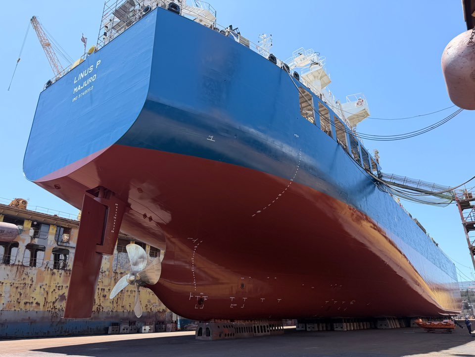 Large blue-and-red cargo ship named Linus P in a shipyard, viewed from beneath its hull with propeller and dry dock equipment visible.