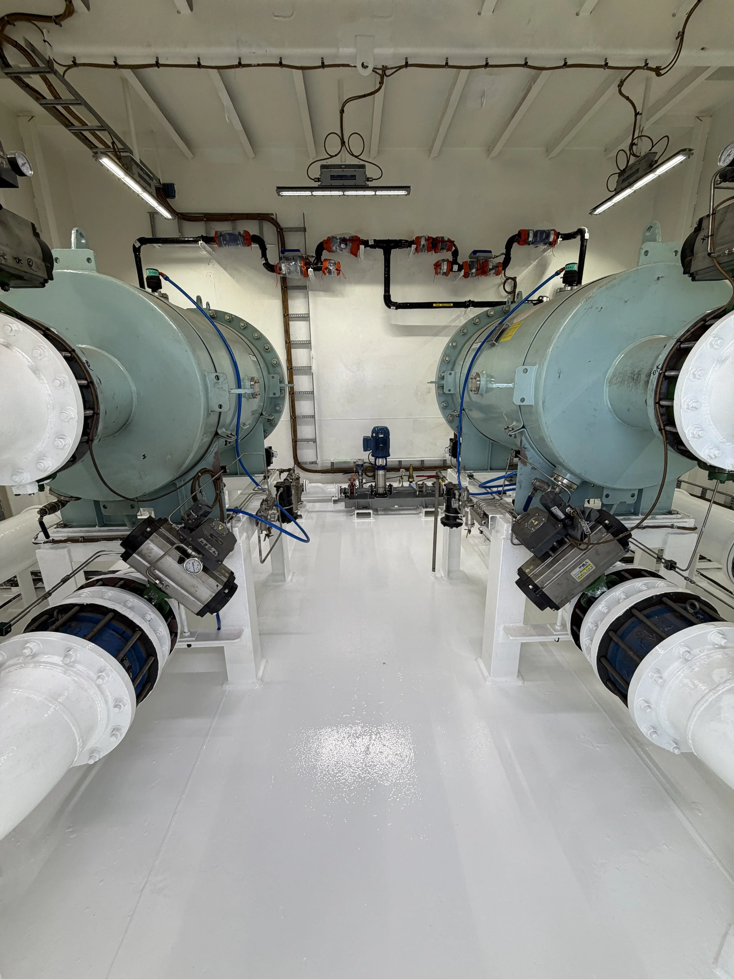Industrial machinery room with large flanged pipes, valves, blue pneumatic tubing, overhead wiring, and fluorescent lights