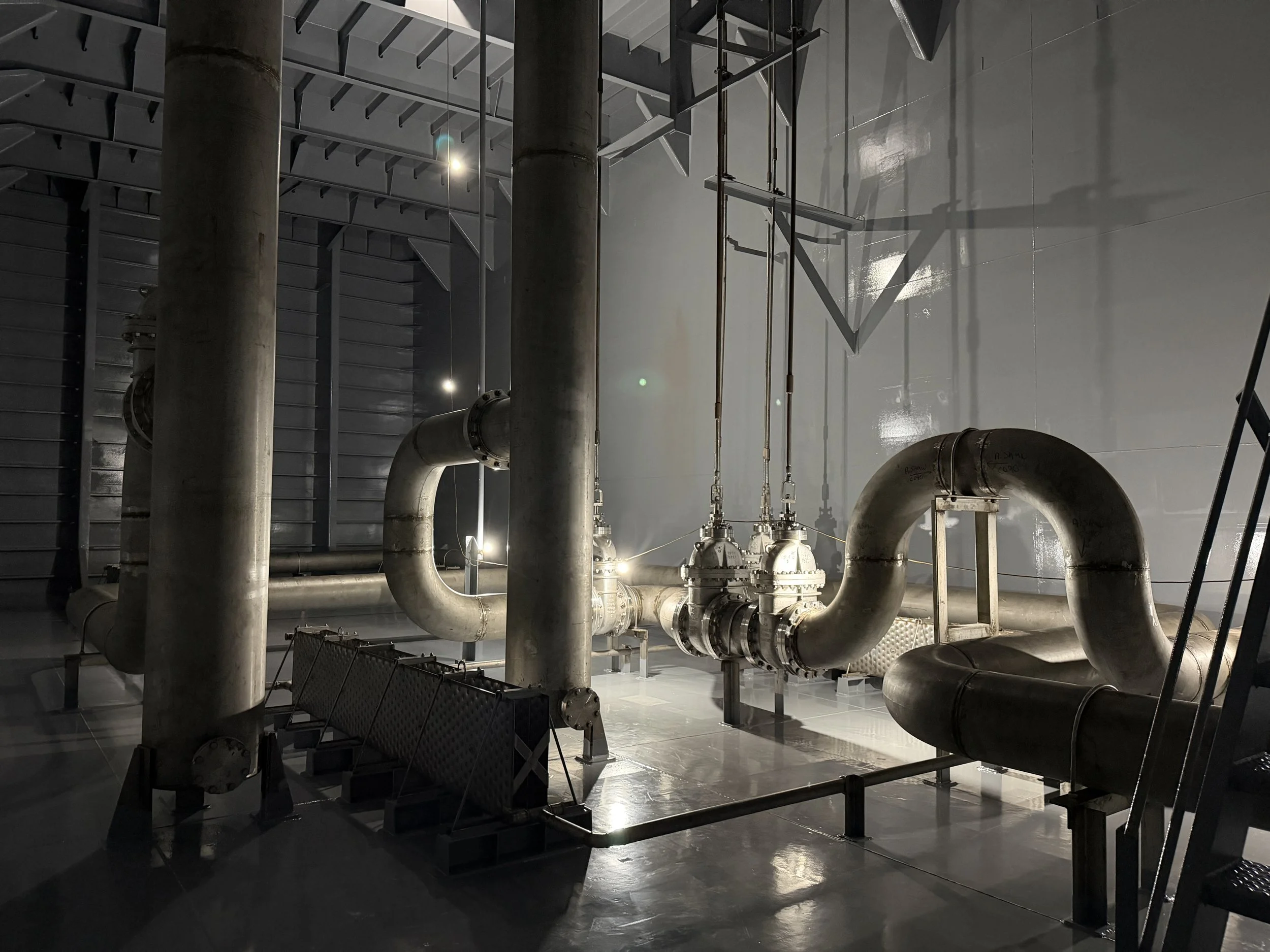 Industrial facility interior with large metal pipes, valves, cylindrical tanks, and structural steel beams