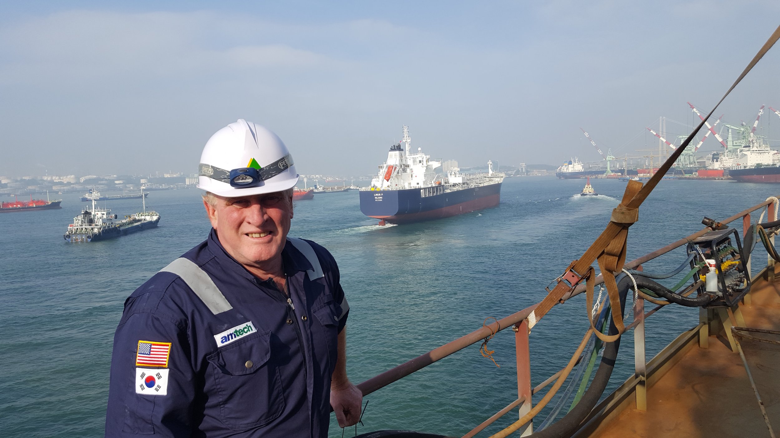 Maritime worker wearing a hard hat and coveralls aboard a ship, with cargo vessels and an industrial harbor in the background.