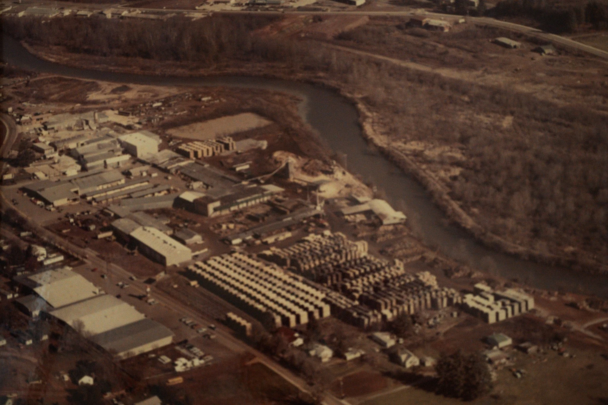 Aerial view of a small industrial area next to a winding river, with various warehouses and buildings surrounded by roads and open land.