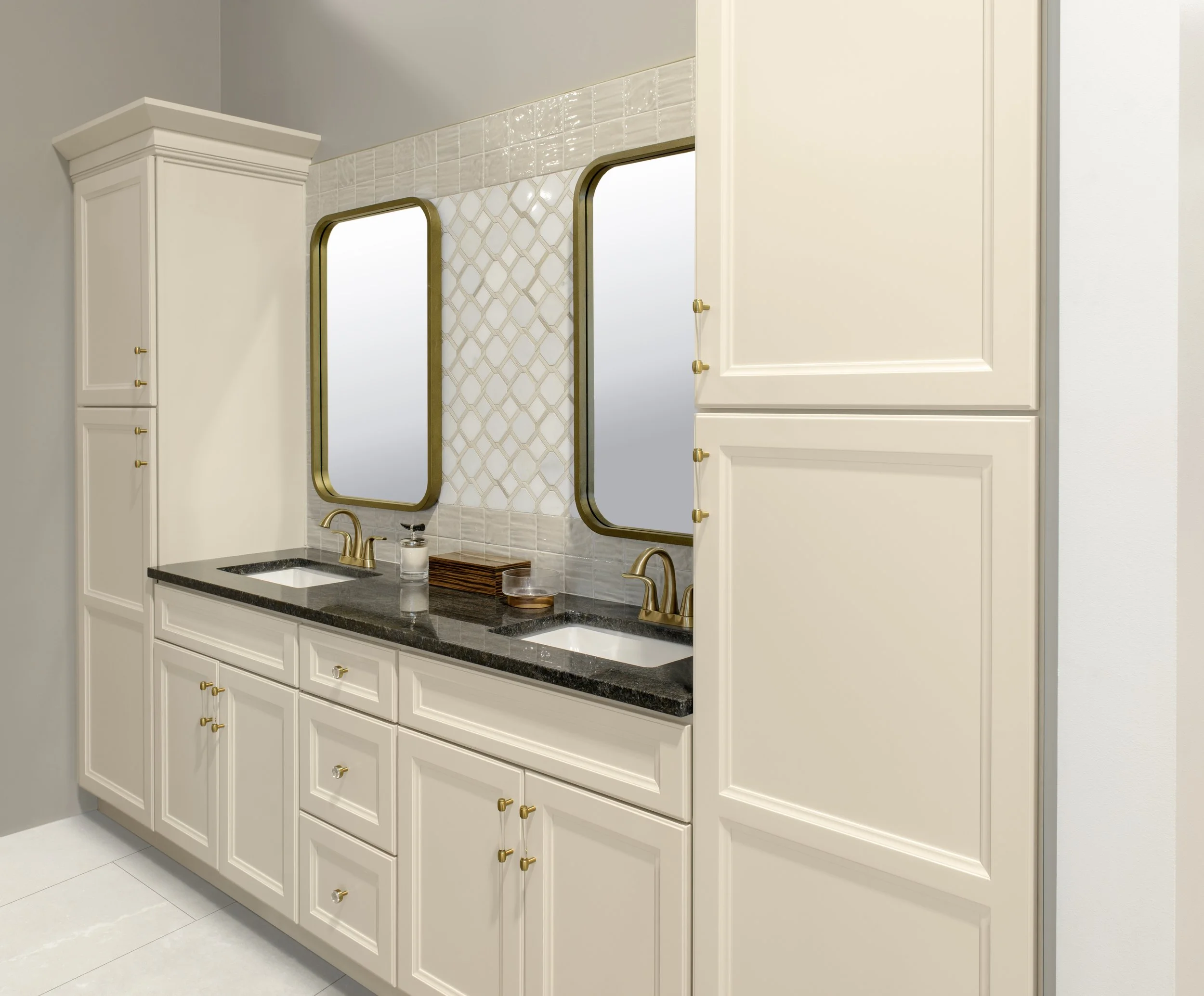A bathroom vanity with white cabinets, gold hardware, a black countertop, a double sink, two mirrors with gold frames, and wall tiles with a beige and white geometric pattern.