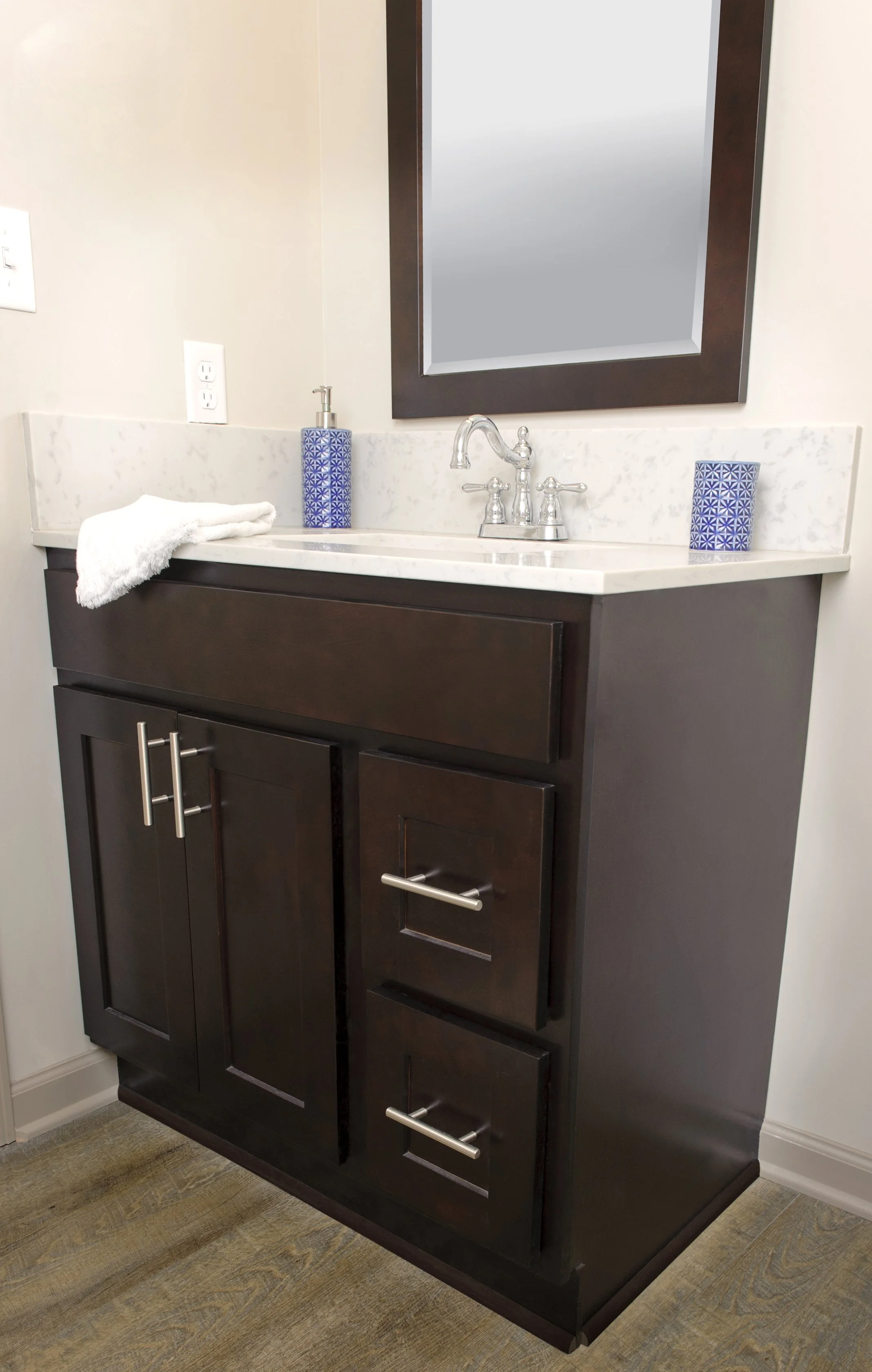 Bathroom vanity with a dark wood cabinet, a white marble countertop, a mirror, a silver faucet, blue patterned soap dispensers, a white towel, and electrical outlets on a beige wall.