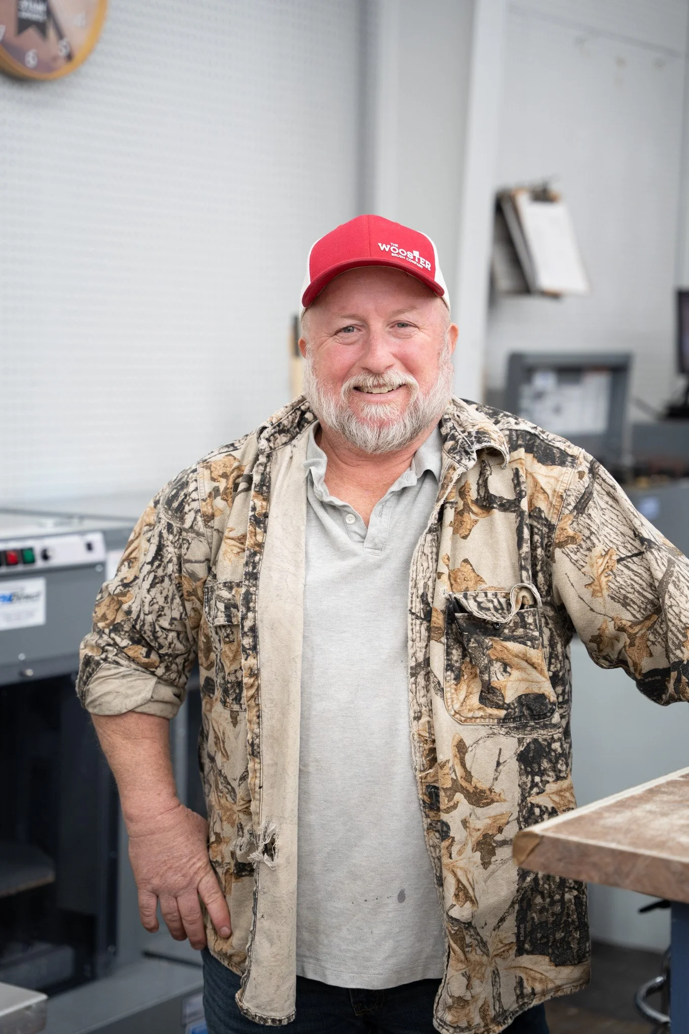 A smiling man with a gray beard and mustache wearing a red hat, camouflage jacket, and gray shirt, standing indoors.