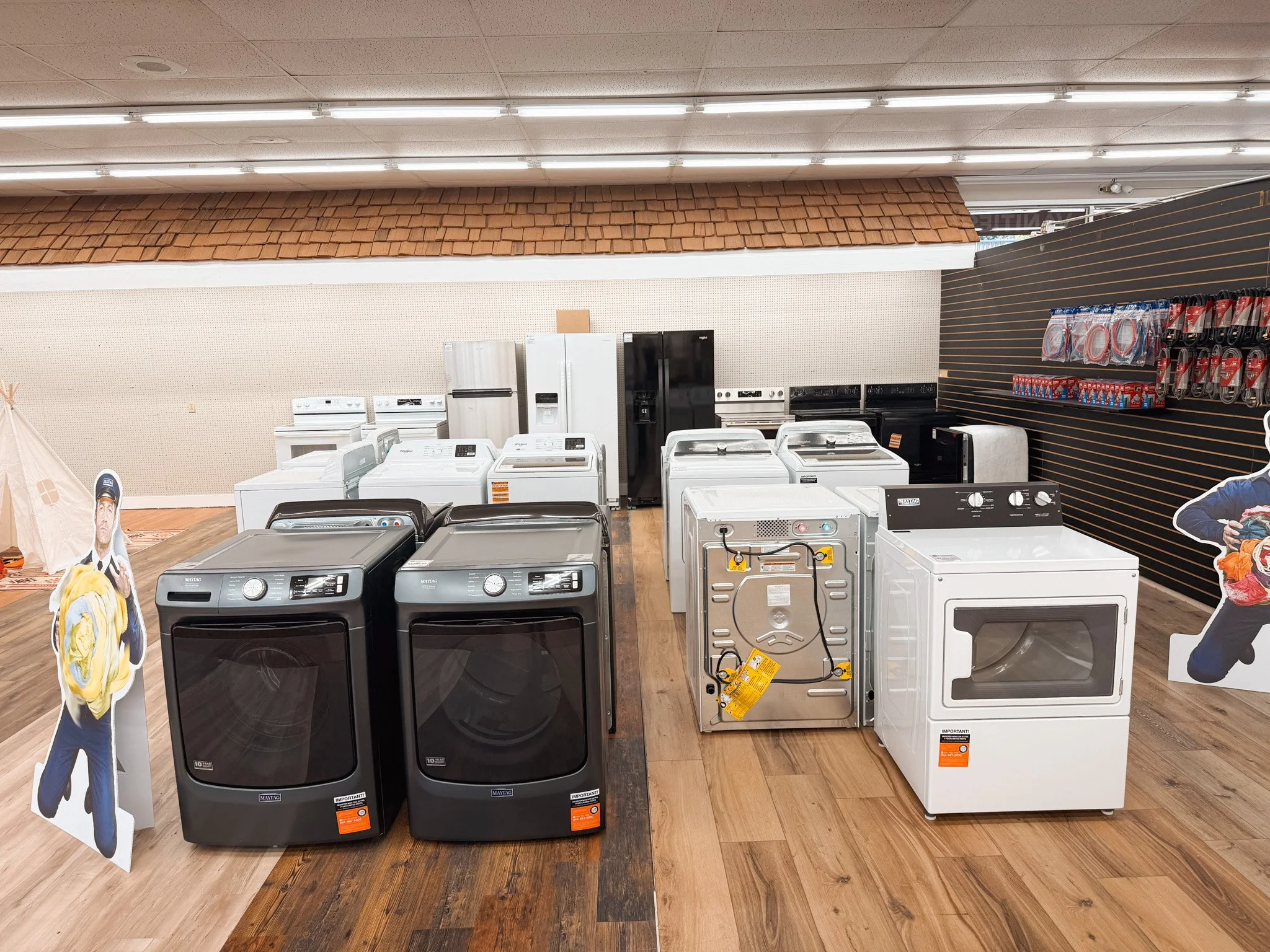 Display of washing machines and dryers in an appliance store with cardboard cutouts of people on either side.