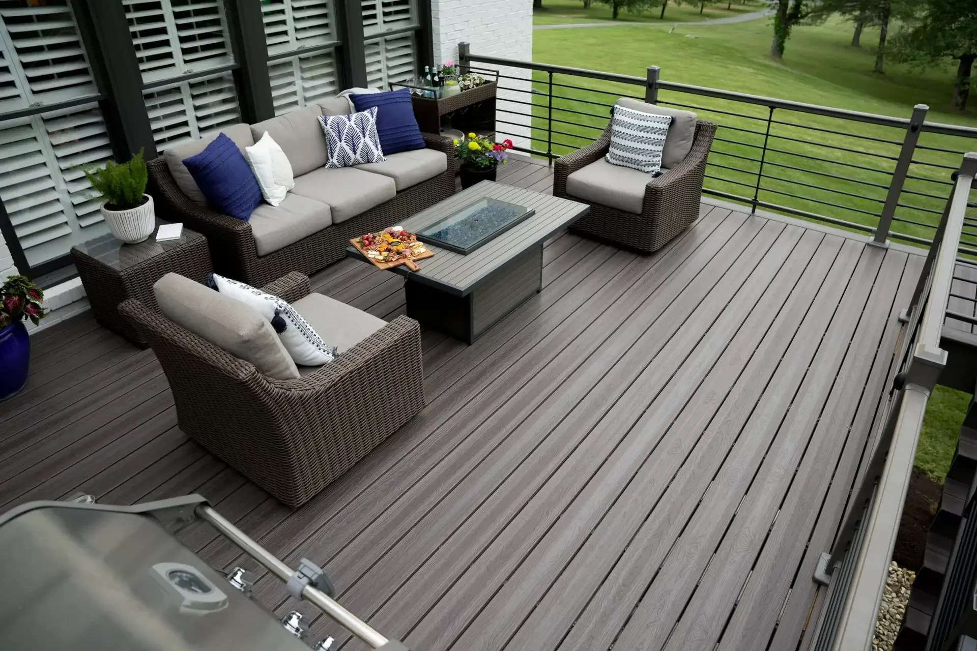 Outdoor wooden deck with wicker furniture including a sofa, two armchairs, a coffee table with decorative tray, potted plants, and pillows, overlooking a green lawn and trees.