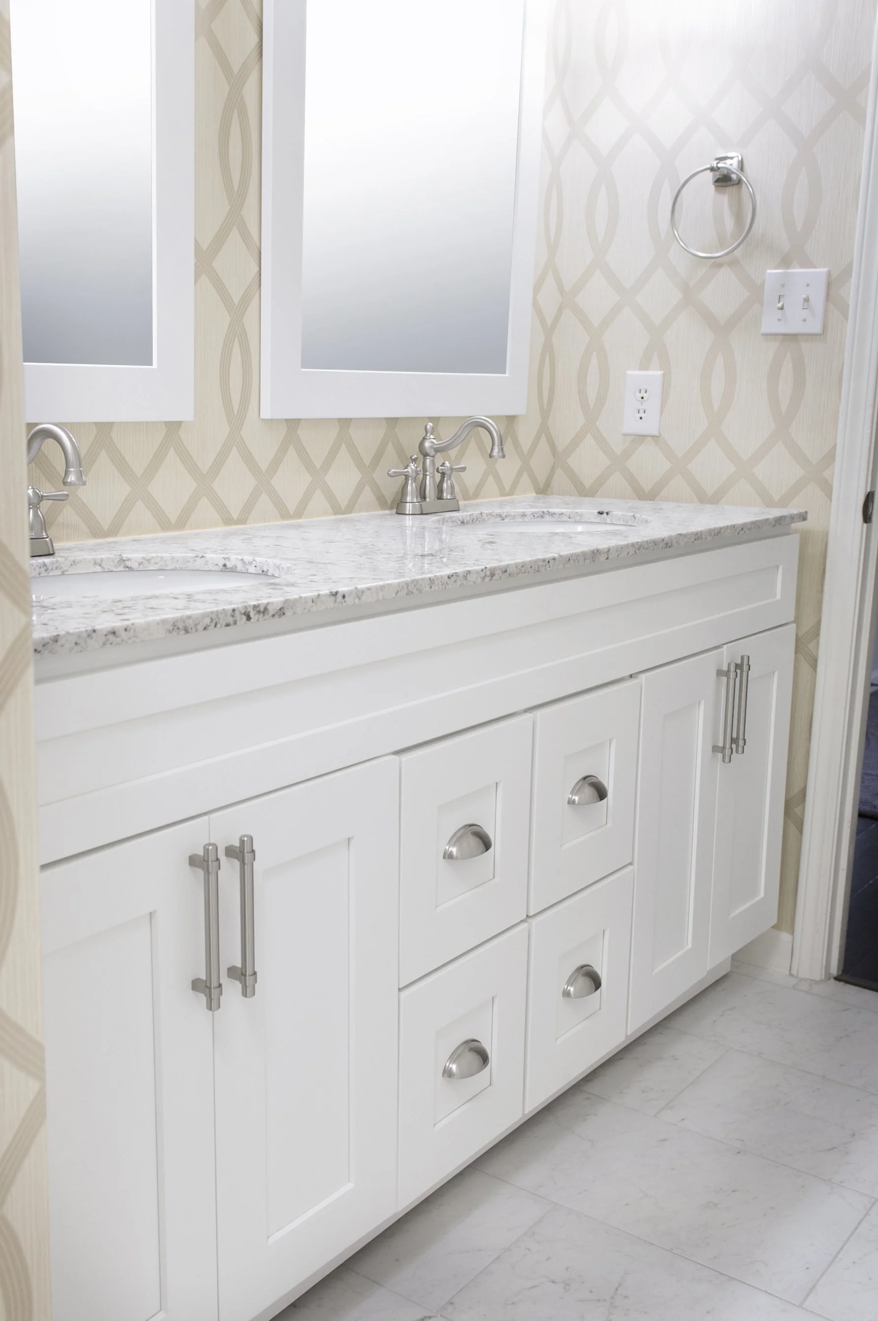 Bathroom double vanity with white cabinets, marble countertop, two sinks, and silver faucets. Two mirrors above the sinks, one towel ring on the right wall, power outlets beside the mirrors, and a cream-colored wallpaper with a geometric pattern.