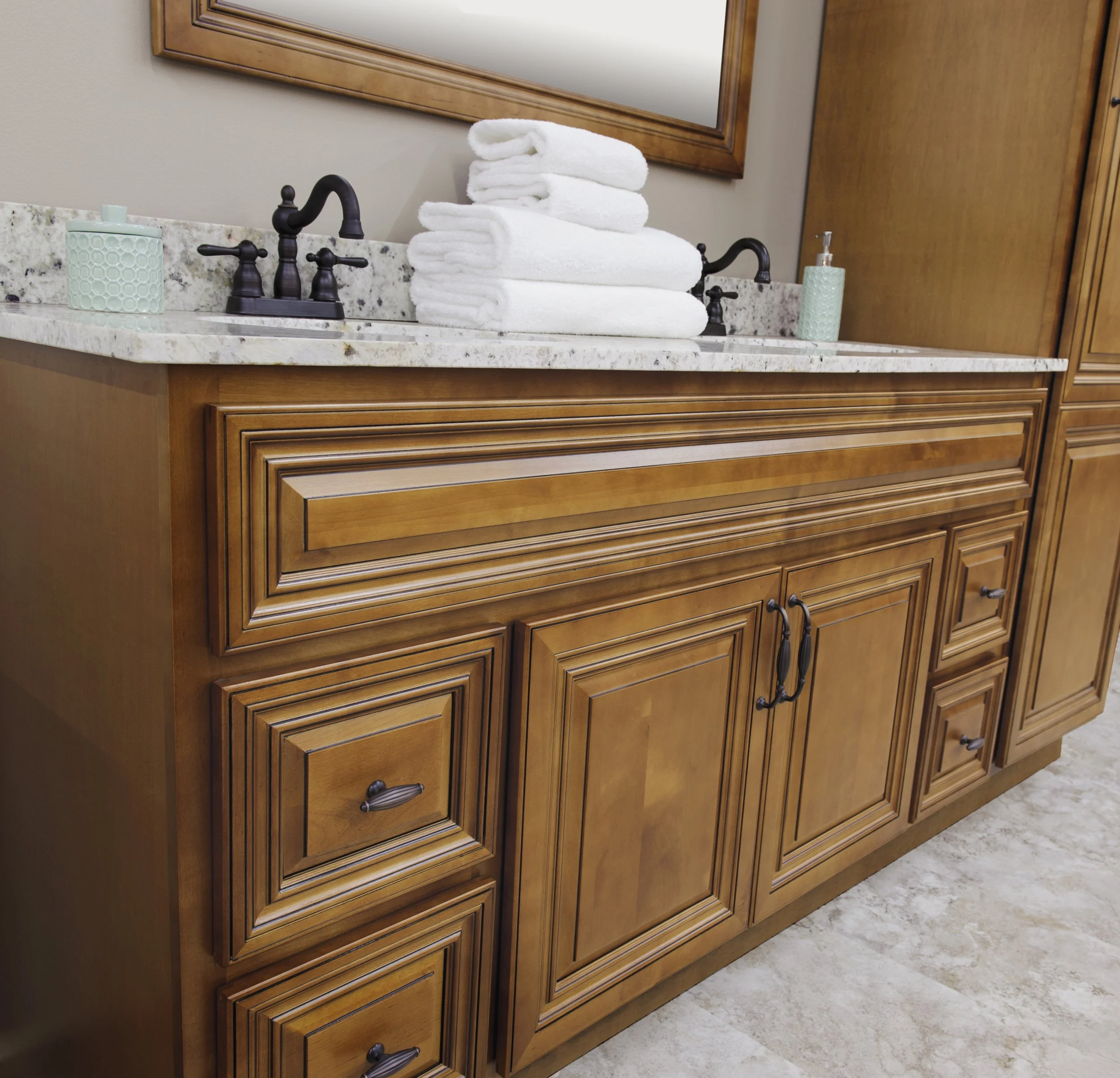 Wooden bathroom vanity with black handles, a marble countertop, a mirror, a stack of folded white towels, a green ceramic soap dispenser, and a green ceramic cup with a toothbrush inside.