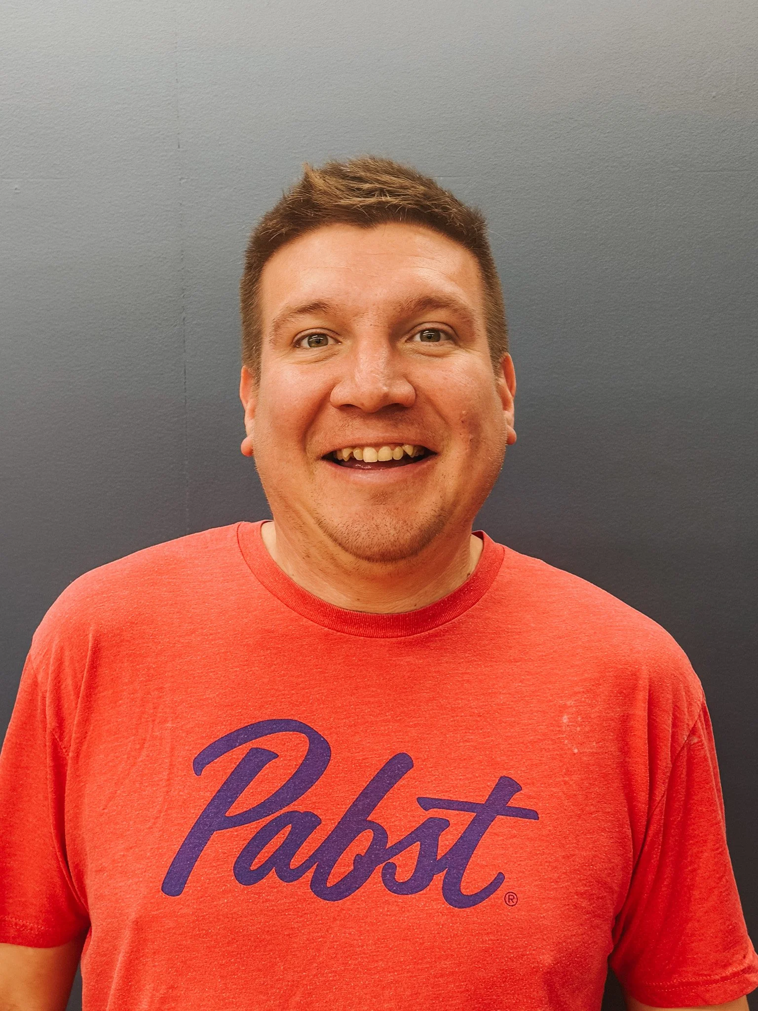 A man smiling, wearing a red T-shirt with the word 'Pabst' written in purple, standing in front of a plain, dark gray background.