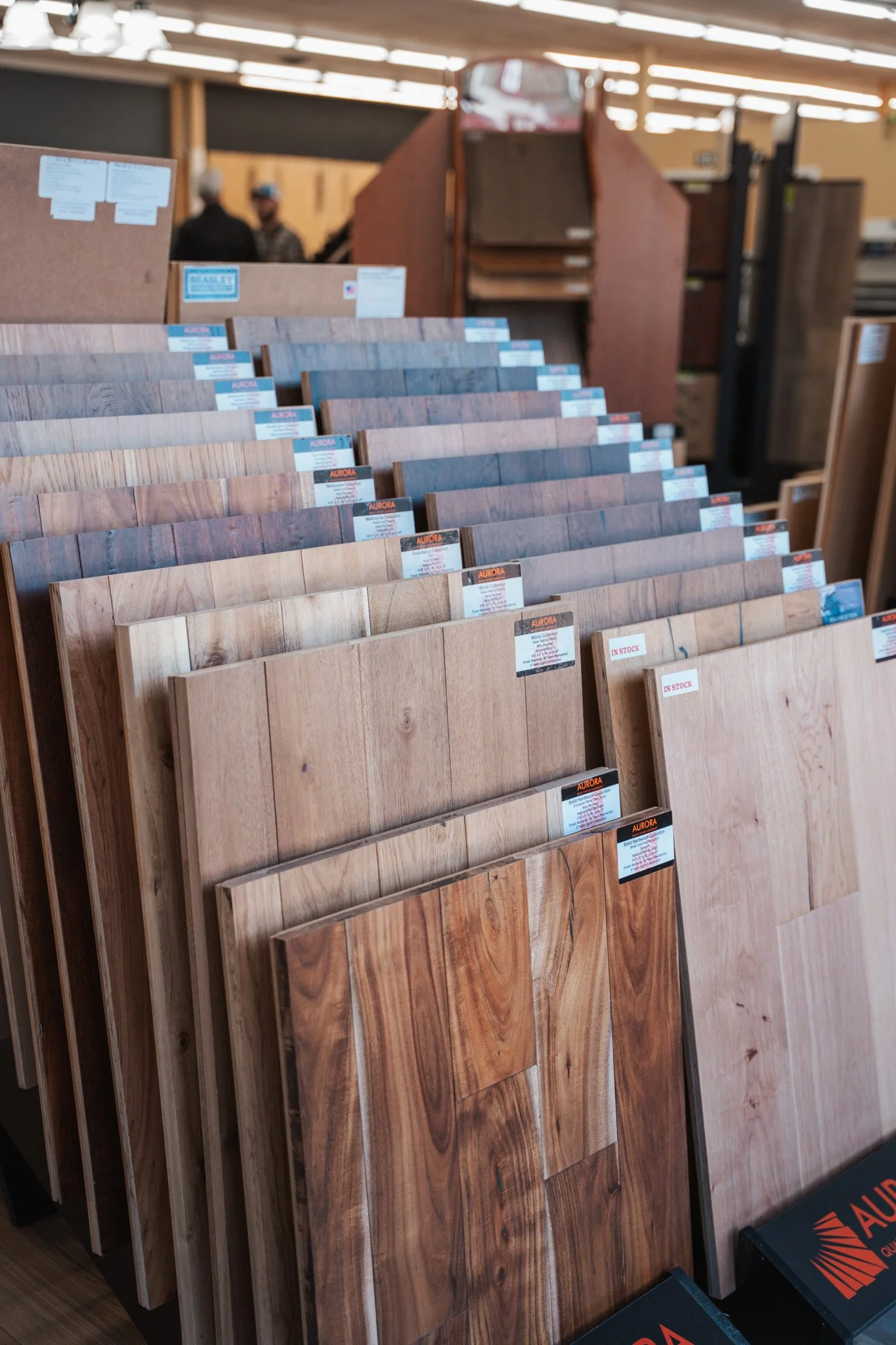Various hardwood flooring samples displayed on racks in a store, with some labeled 'In Stock' and brand 'Aurora' visible.