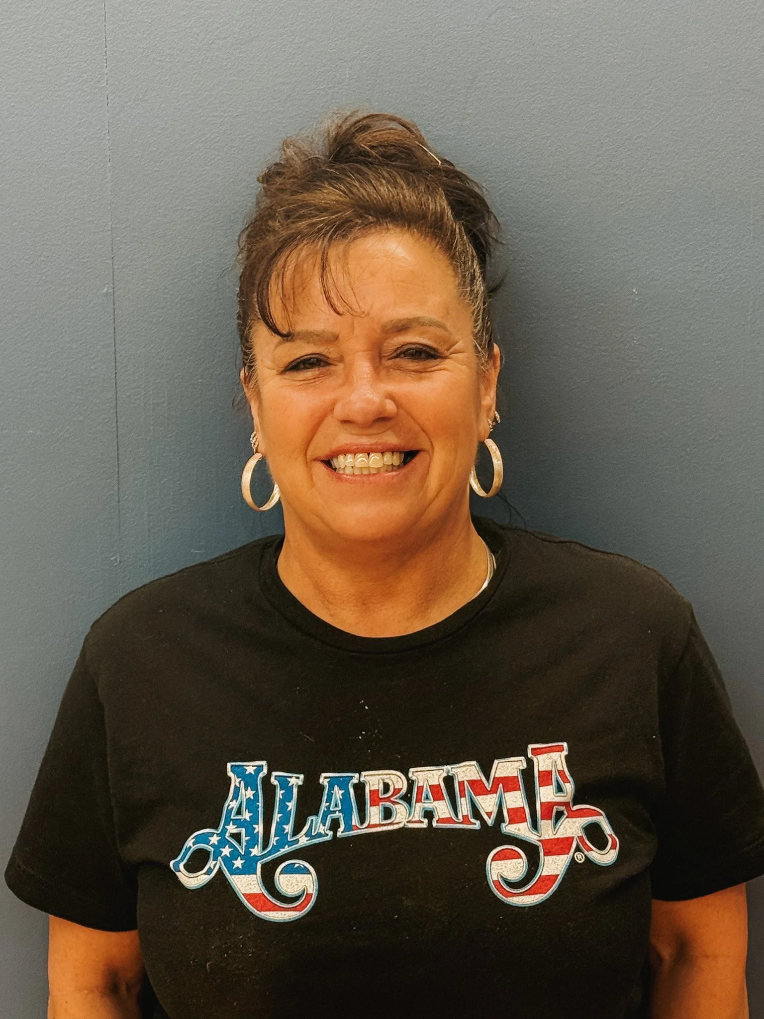 Smile woman wearing a black T-shirt with 'ALABAMA' written in patriotic colors, standing against a blue wall.