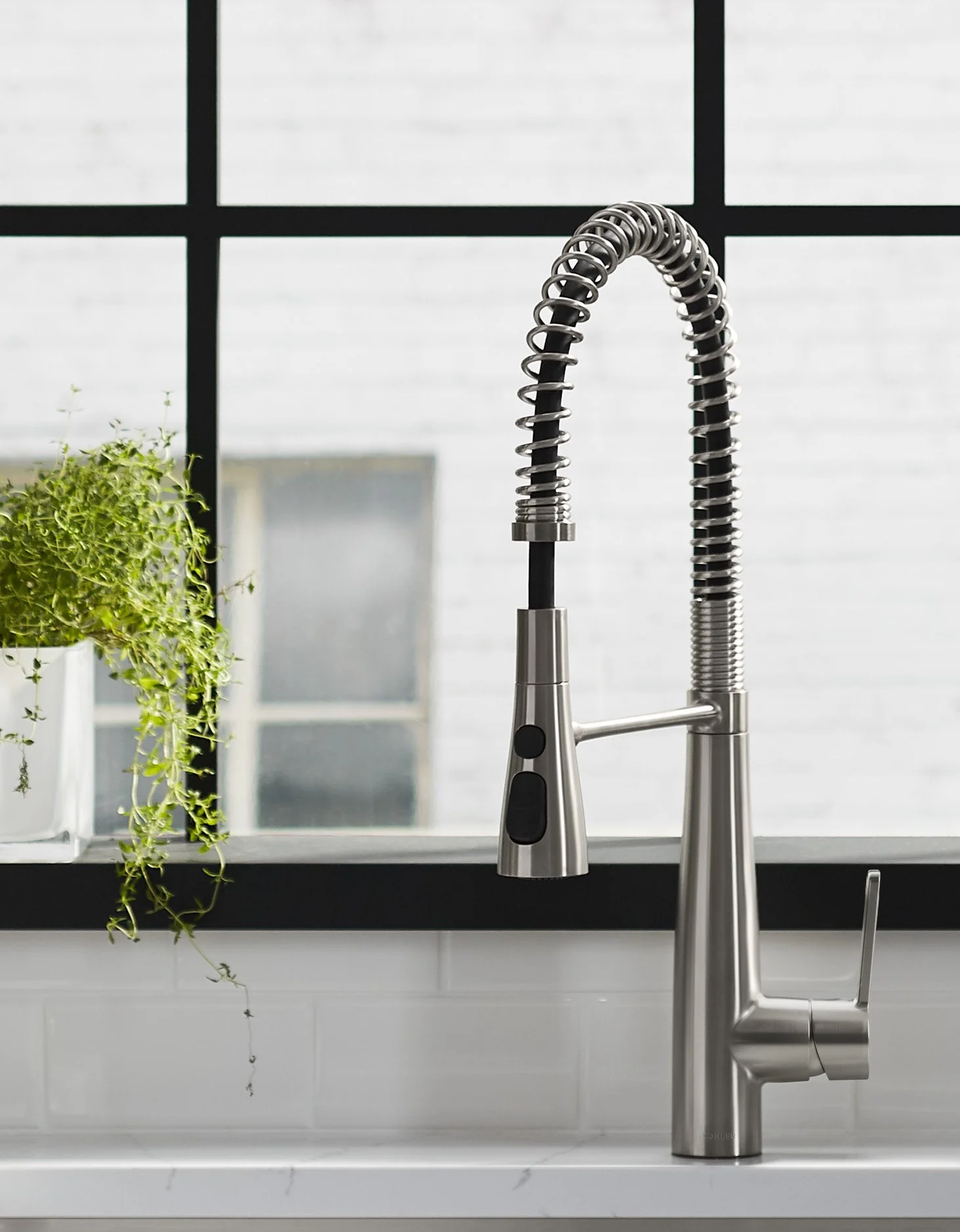 A modern kitchen sink with a tall, curved, stainless steel faucet next to a potted green herb plant on a white countertop, with a large window in the background.