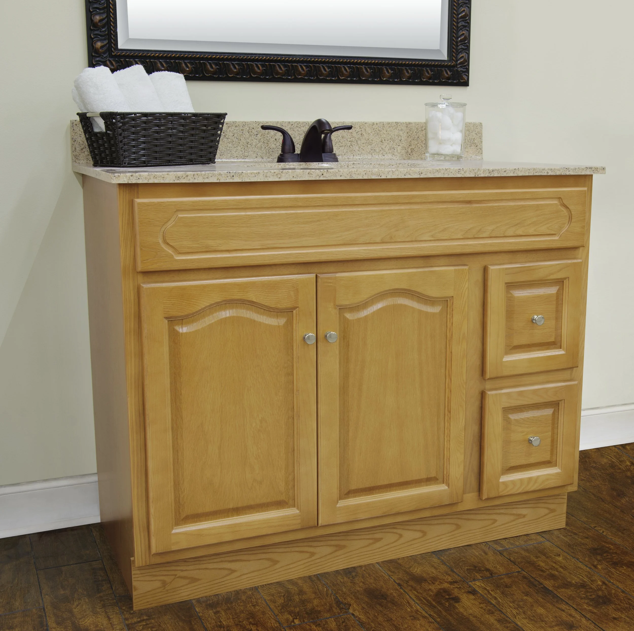 Wooden bathroom vanity with a granite countertop, black faucet fixtures, a basket of white towels, and a glass jar filled with cotton balls, with a mirror above.