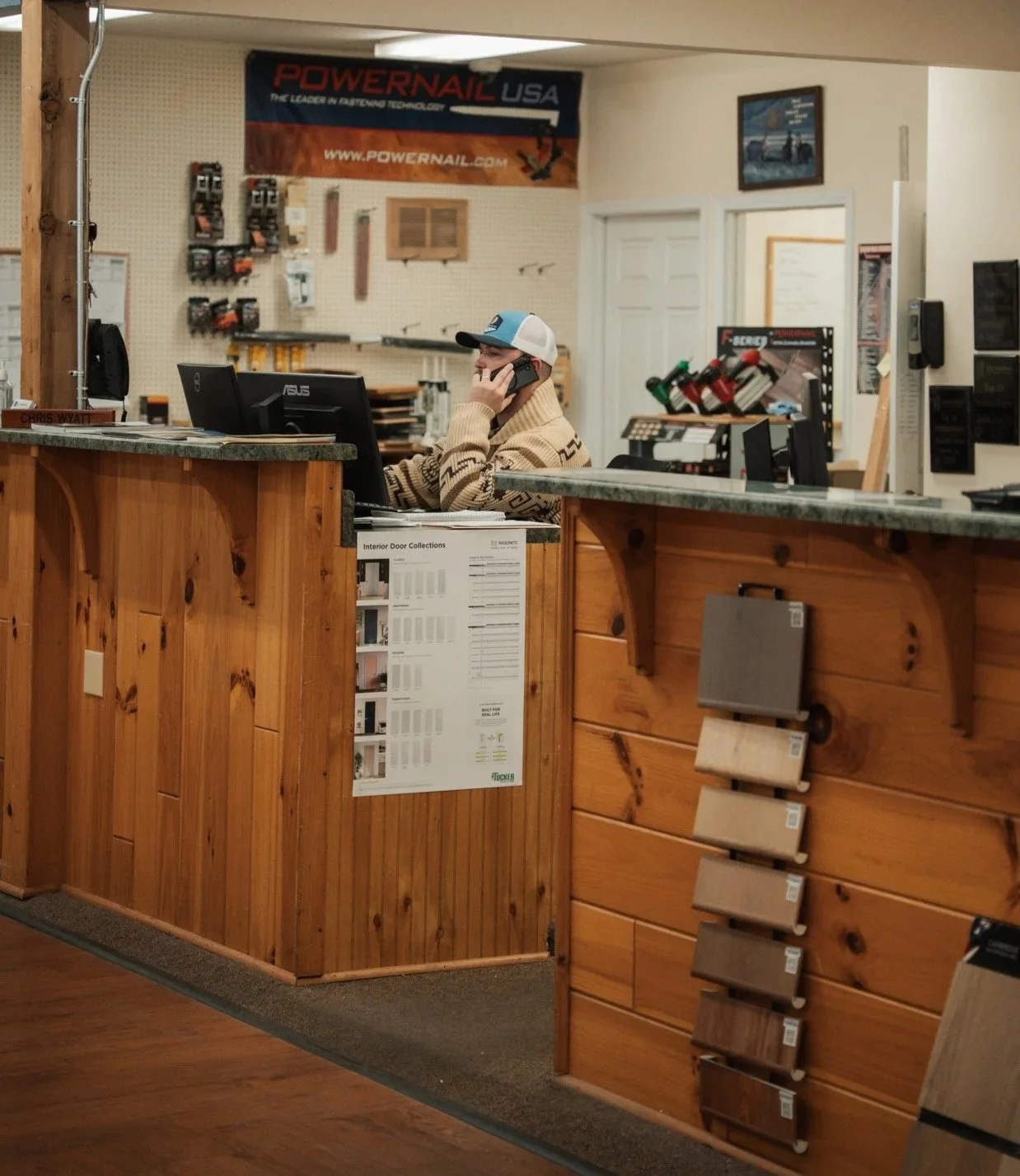 Interior of a hardware store with a cashier wearing a beige sweater and a blue cap, talking on the phone behind the counter made of wood, with various hardware products and samples displayed around.