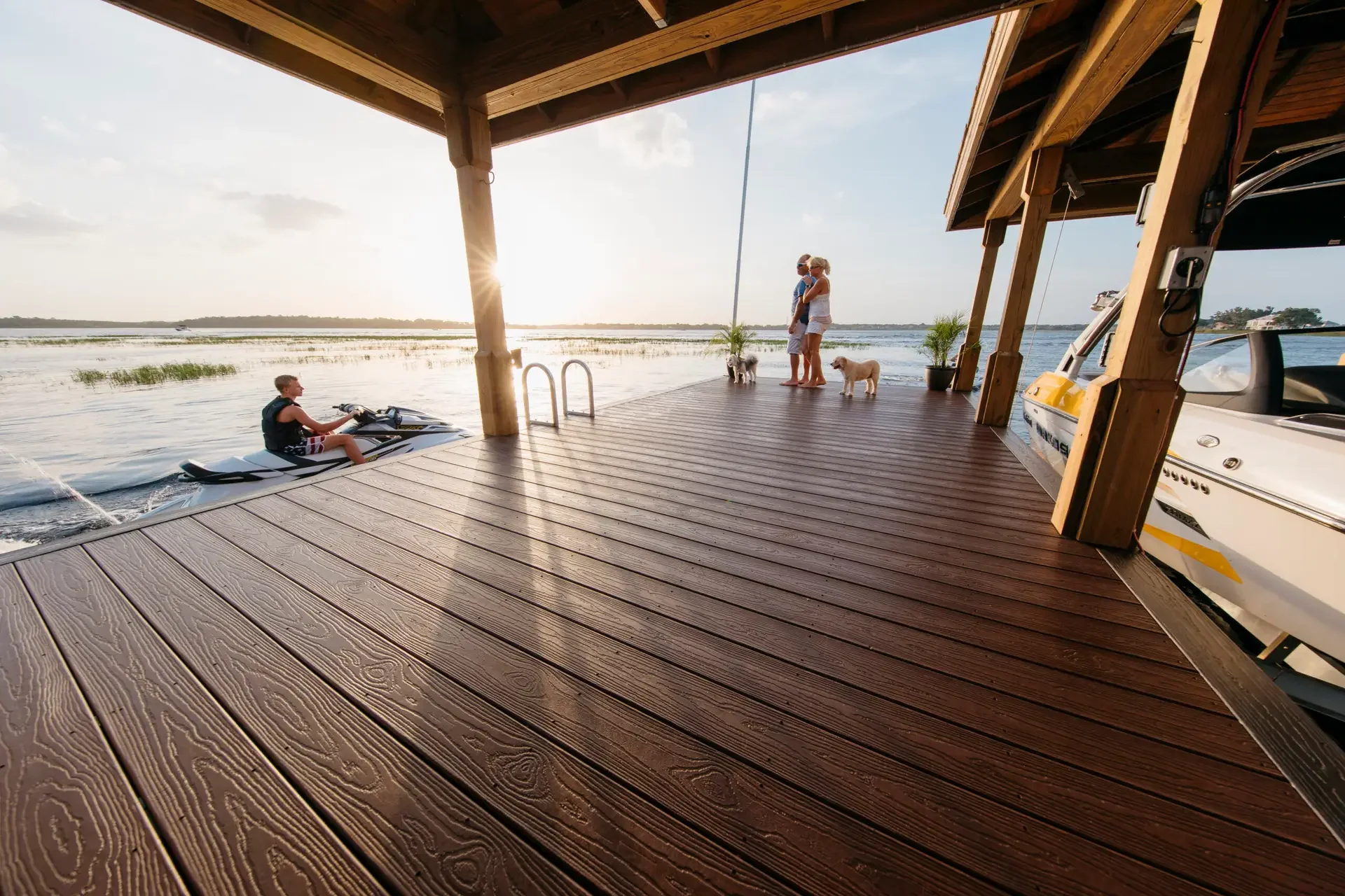 A family with dogs enjoying a sunset on a wooden dock, with a person riding a jet ski and a boat docked nearby.