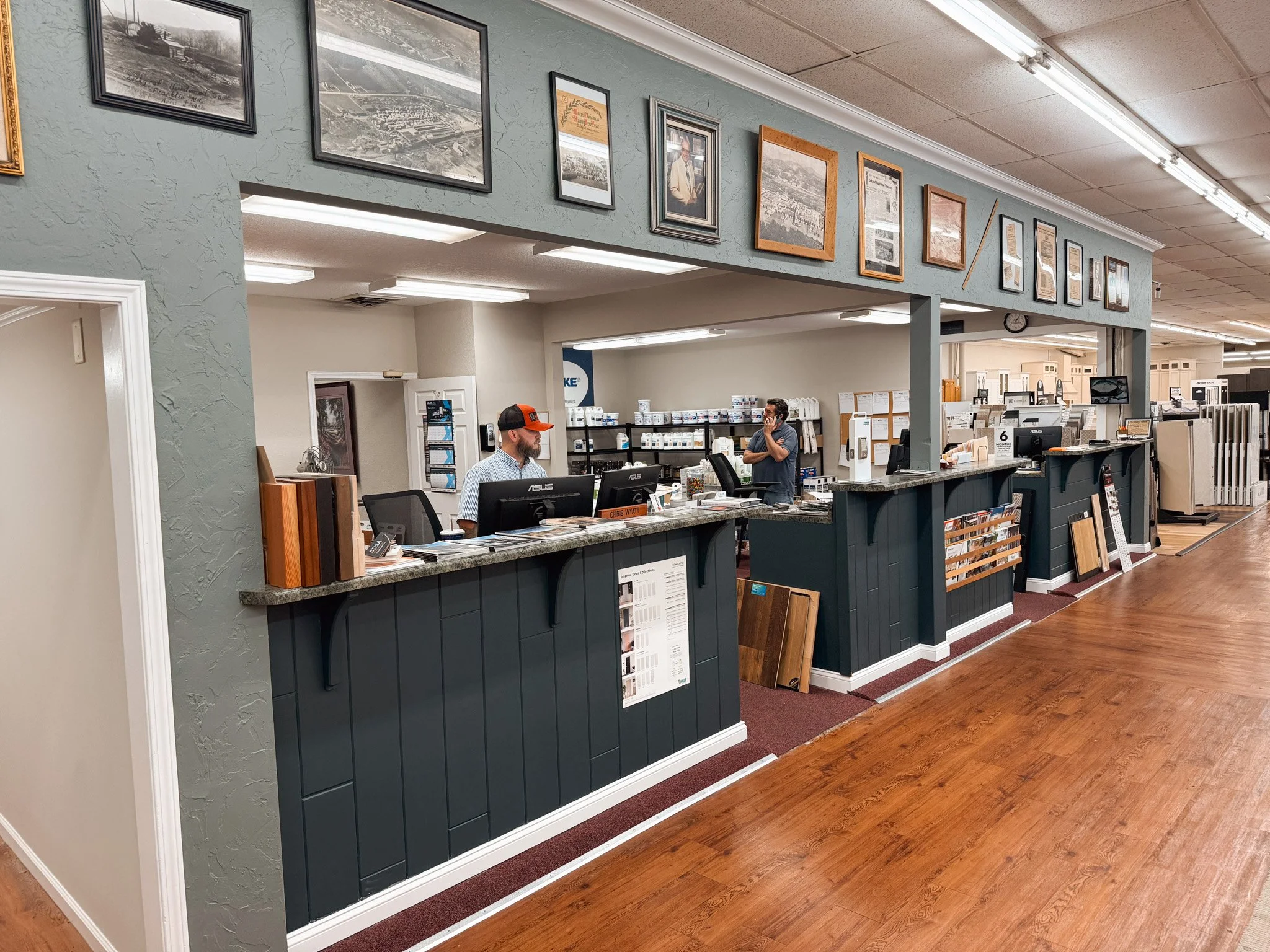 Inside an office or retail store, featuring a checkout counter with two men, one working on a computer and the other on the phone. The counter is decorated with brochures and sample materials, and there are framed pictures and newspapers on the wall above.