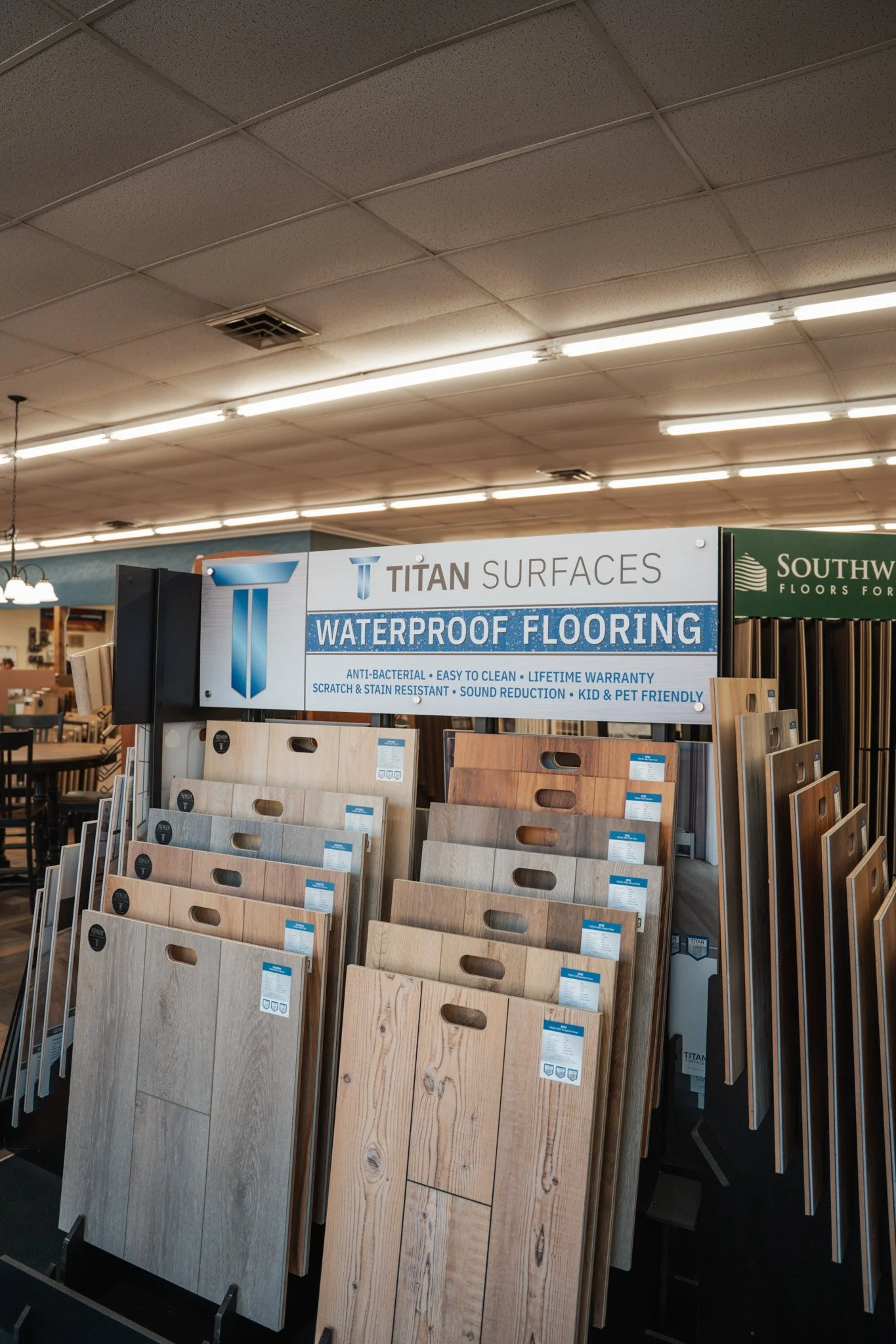 Display of waterproof flooring samples in a hardware store, with various wood-like textures and colors arranged on racks beneath a sign that reads "Titan Surfaces Waterproof Flooring."