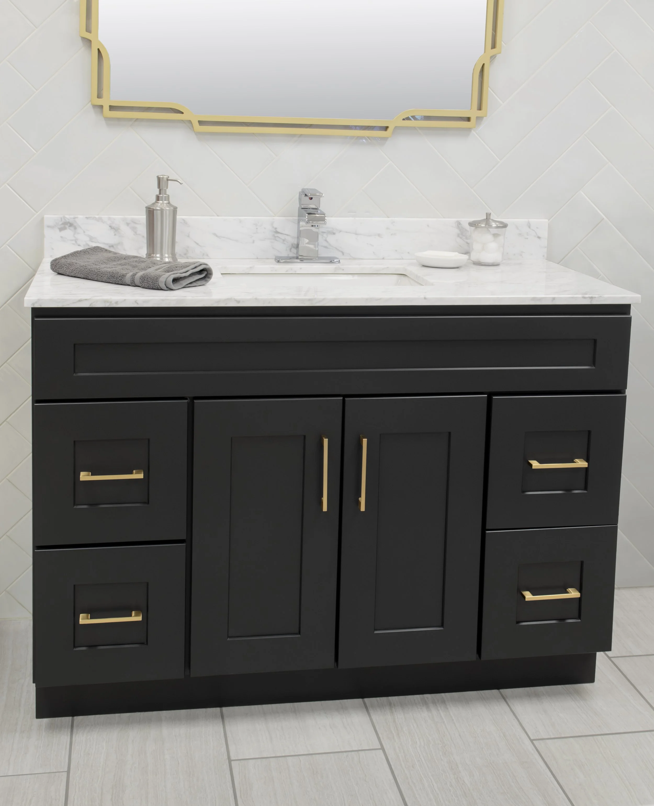 A black bathroom vanity with a marble countertop, a silver soap dispenser, a gray towel, a small white bowl, and a jar of cotton balls. A gold-framed mirror hangs above the sink, and the wall behind is tiled with beige diamond-shaped tiles.