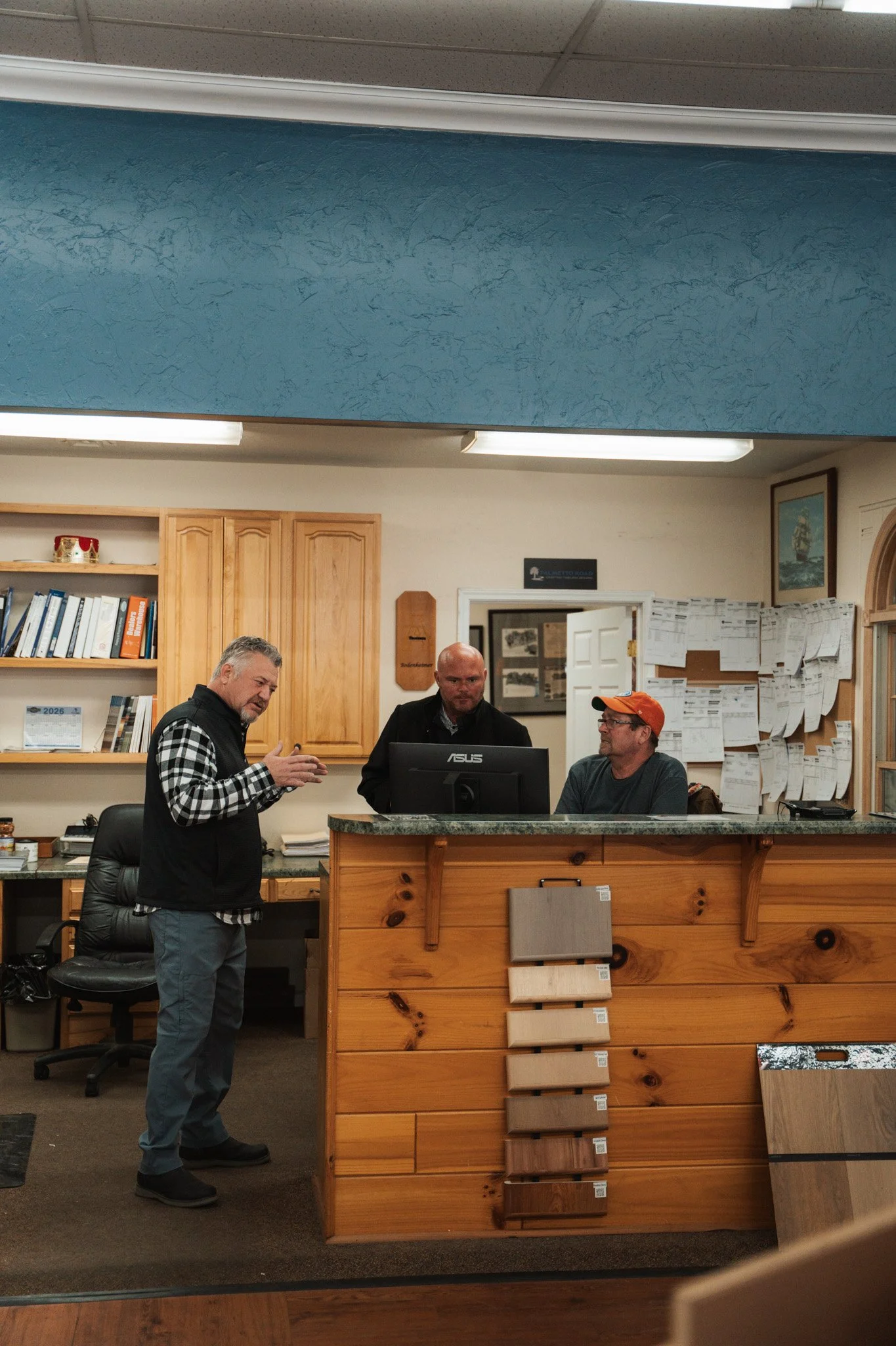 Three men are having a conversation at a reception desk in an office or store. One person is standing, and two are seated behind the desk. The desk is made of wood with samples of different wood finishes displayed on it. Bookshelves, posters, and papers are visible in the background.
