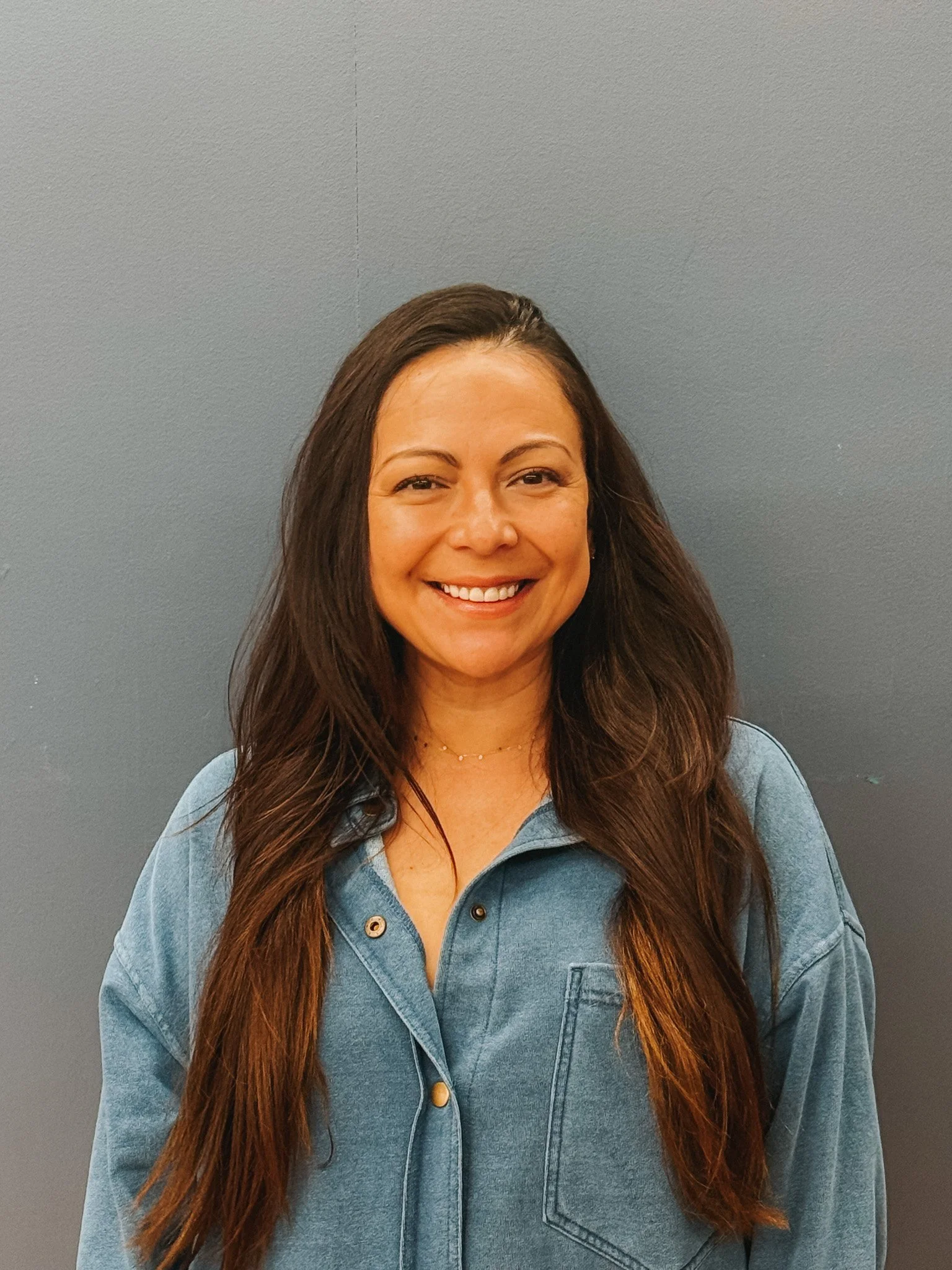 A smiling woman with long dark hair wearing a light blue denim jacket stands against a plain gray wall.