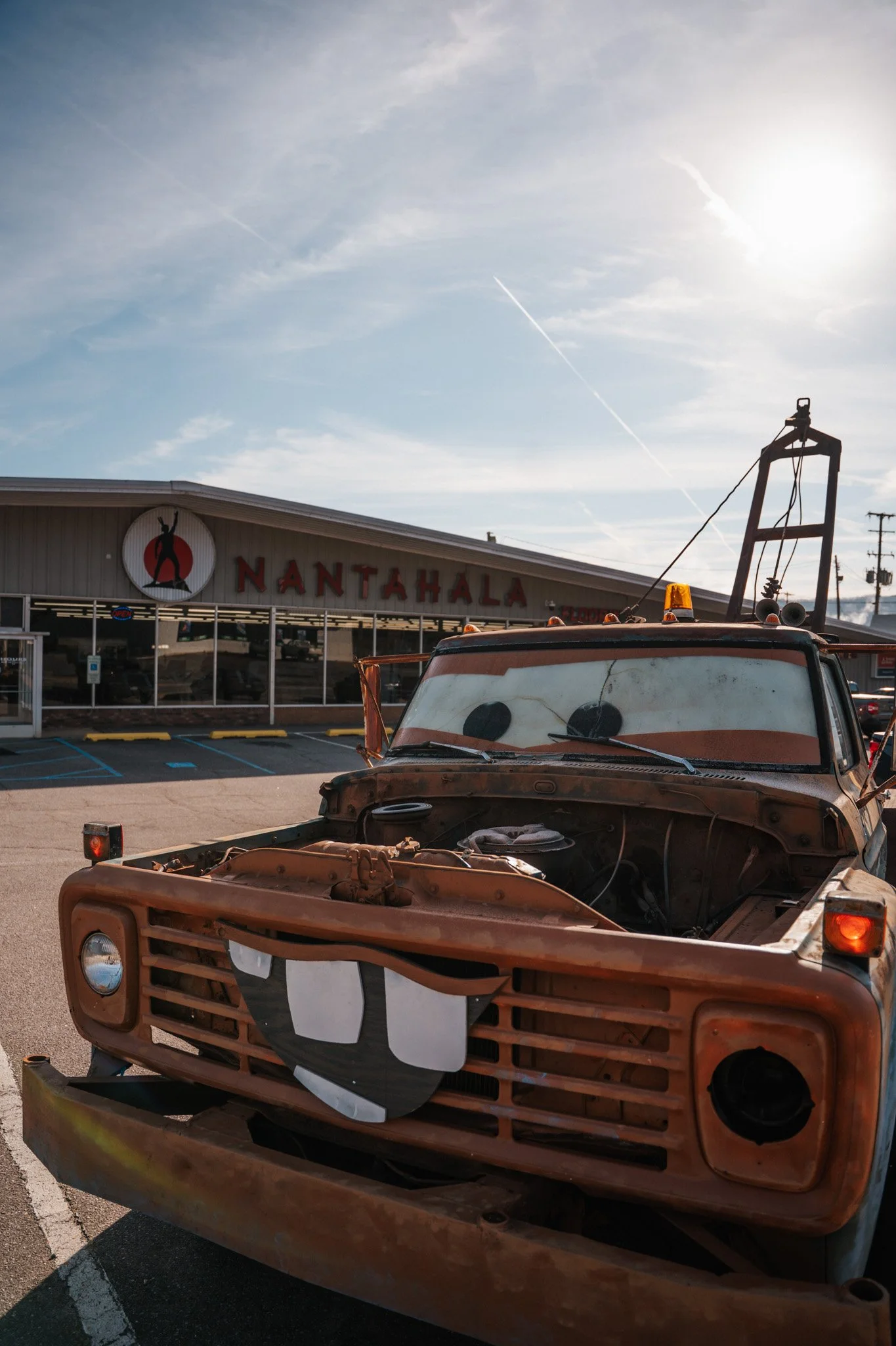 An old, rusty truck with a painted face on the front, parked in front of a NANTAHALA store with a logo of a person dancing, under a partly cloudy sky with the sun shining.