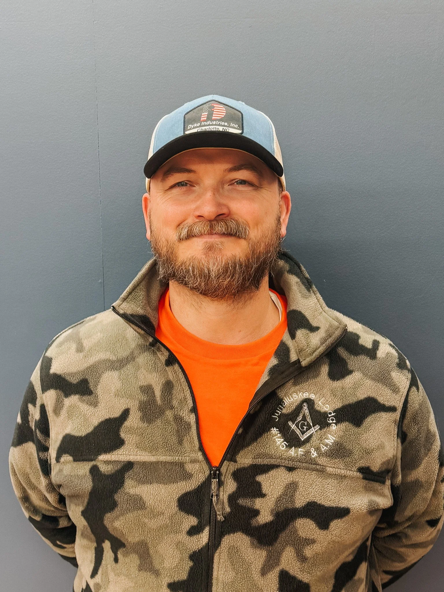 A man with a beard and mustache, wearing a blue and gray cap with a logo, an orange T-shirt, and a camouflage fleece jacket, standing in front of a plain gray wall.