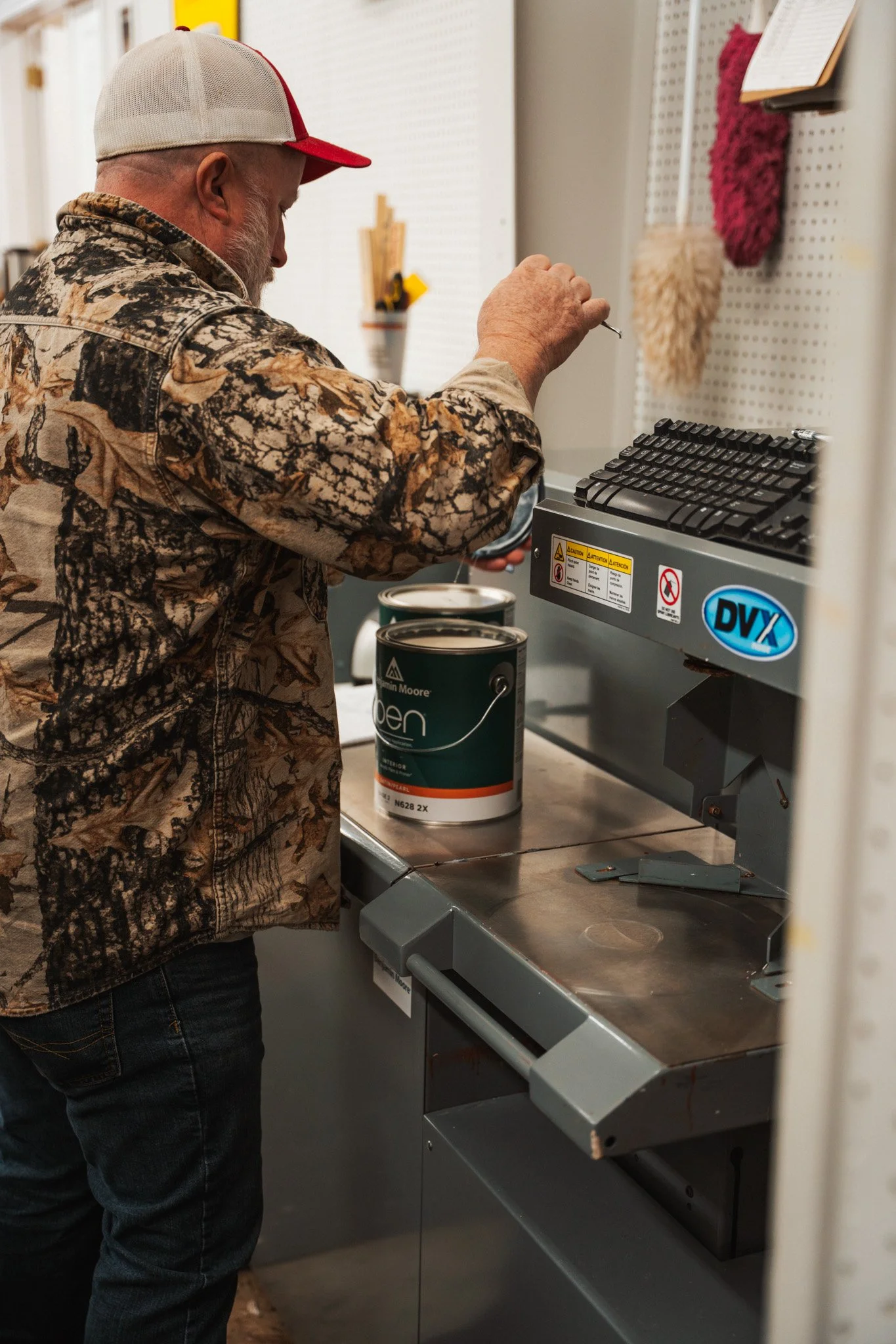 Man in camouflage jacket and cap using a machine at a paint store, with cans of paint on the counter.