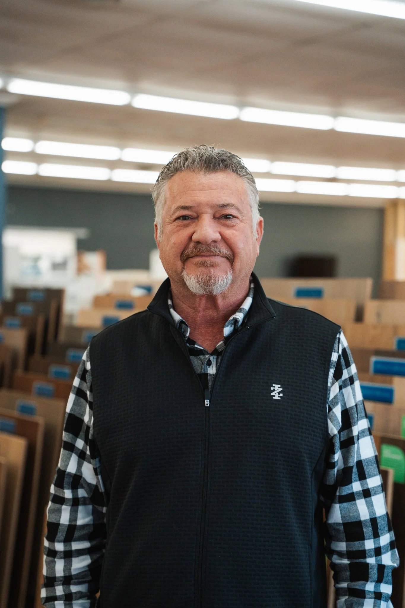 Older man with gray hair and a beard smiling, standing in a store with wood panels and blue stickers on the shelves in the background.