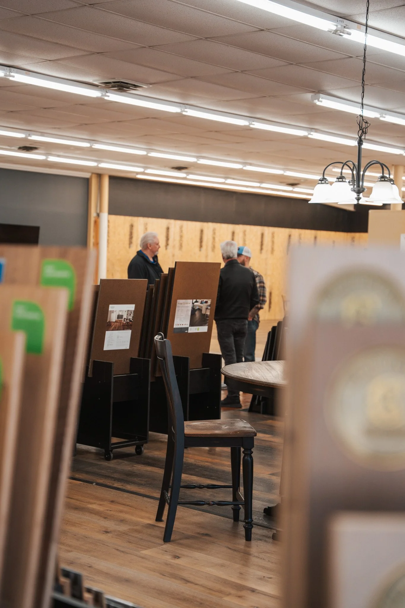 Interior of a store with people shopping, displaying flooring samples, chairs, and lighting fixtures overhead.